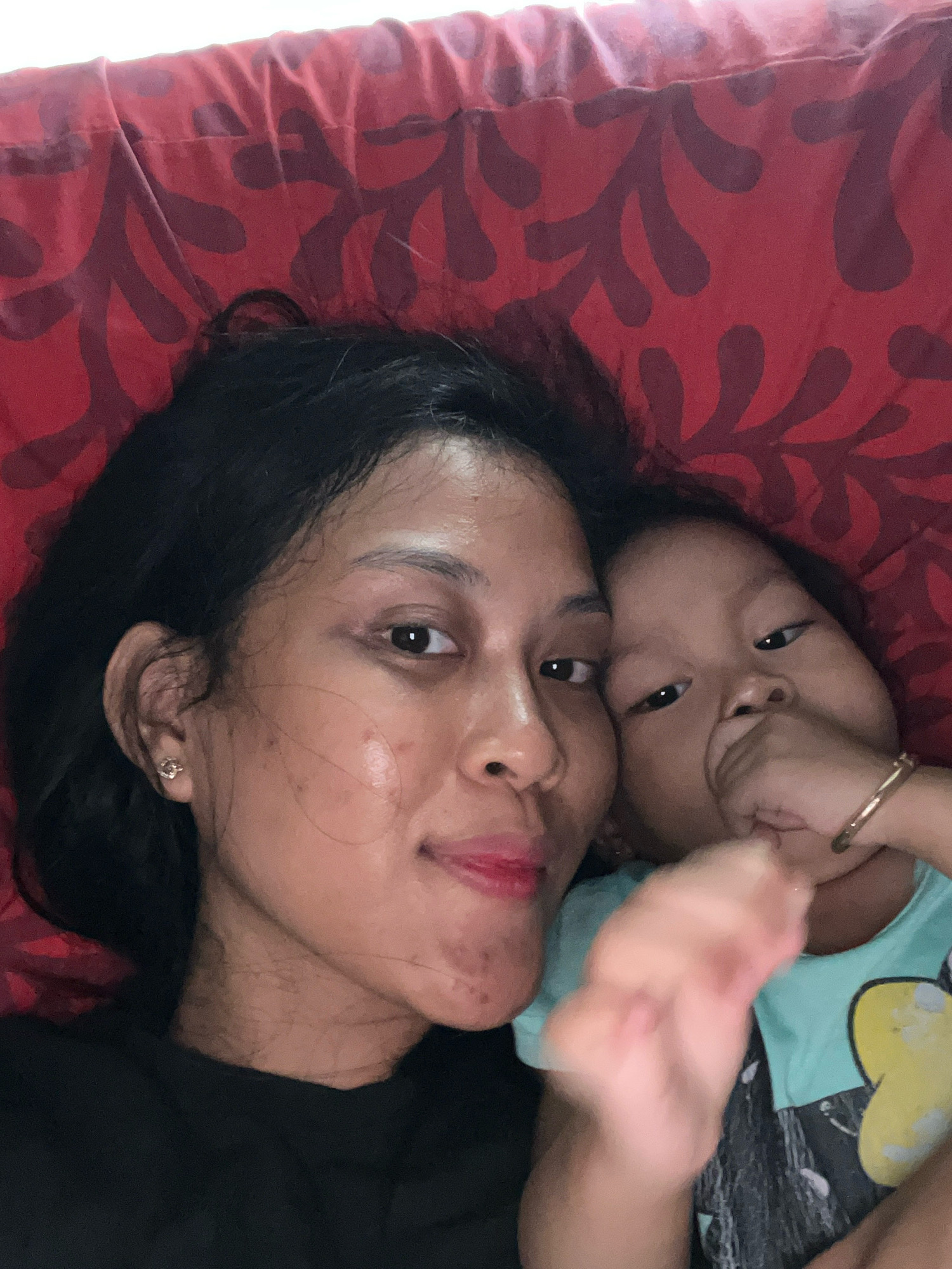 A close-up, overhead, selfie-style photograph shows two individuals, an adult and a child, lying down on a red surface with a dark pattern. The adult, on the left, has dark hair and is wearing a black shirt. They are looking towards the camera with their right eye visible and their lips slightly parted, revealing red lipstick. A small, gold flower-shaped earring is visible in their left ear. The child, on the right, is facing the camera with their mouth partially covered by their right hand. They have dark hair and are wearing a teal shirt with a yellow Mickey Mouse design. A thin gold bangle is on their right wrist. The background is a red fabric with a repeating dark pattern of organic shapes. The overall impression is an intimate moment captured by the individuals themselves. The image was taken in Kecamatan Pati, Indonesia.