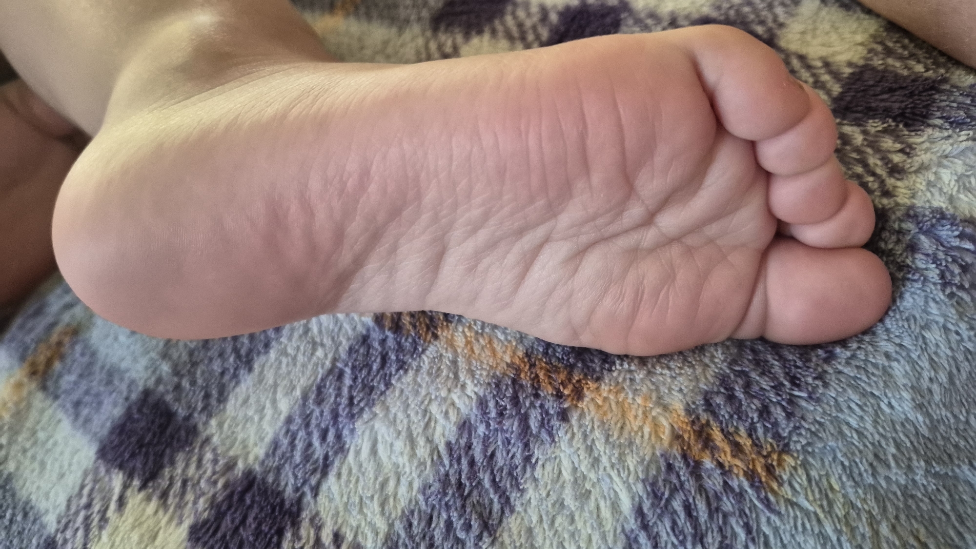 A close-up, overhead shot showcases the sole of a person's foot resting on a textured, multi-colored blanket. The blanket features a pattern of horizontal stripes in shades of purple, blue, and orange, with a fuzzy texture. The foot is pinkish in tone, with visible creases and texture on the sole, particularly around the arch and toes. The heel is rounded, and the toes are curled slightly. The lighting suggests an indoor setting. The location provided is Красназвёздаўскі сельскі Савет, Belarus, but there are no visual cues in the image to confirm this specific location.