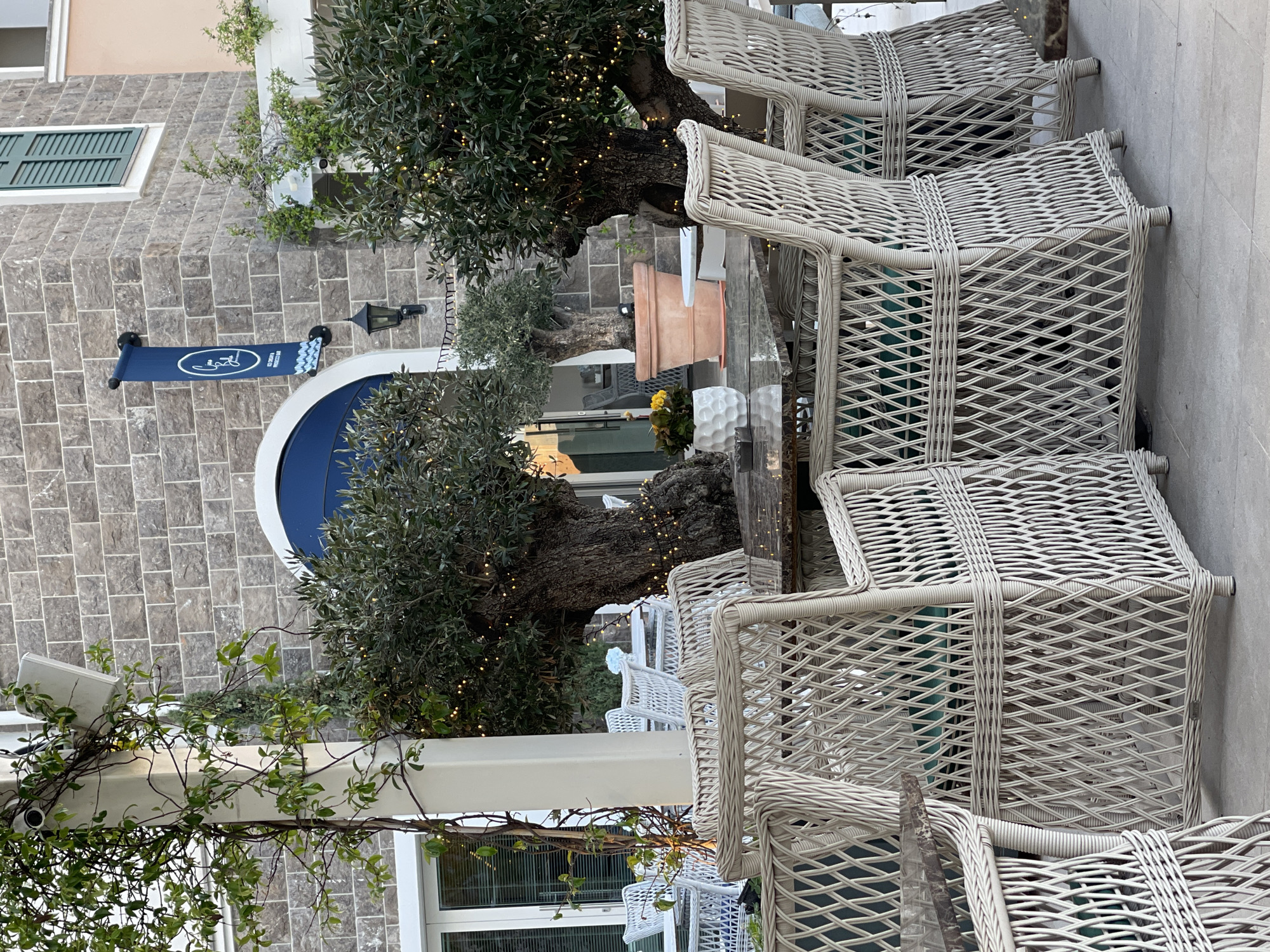 Outdoor patio scene in Radovići, Montenegro. Features wicker chairs around tables, trees with lights, and a building with a blue arched doorway. A sign is seen outside the building, indicating the location.