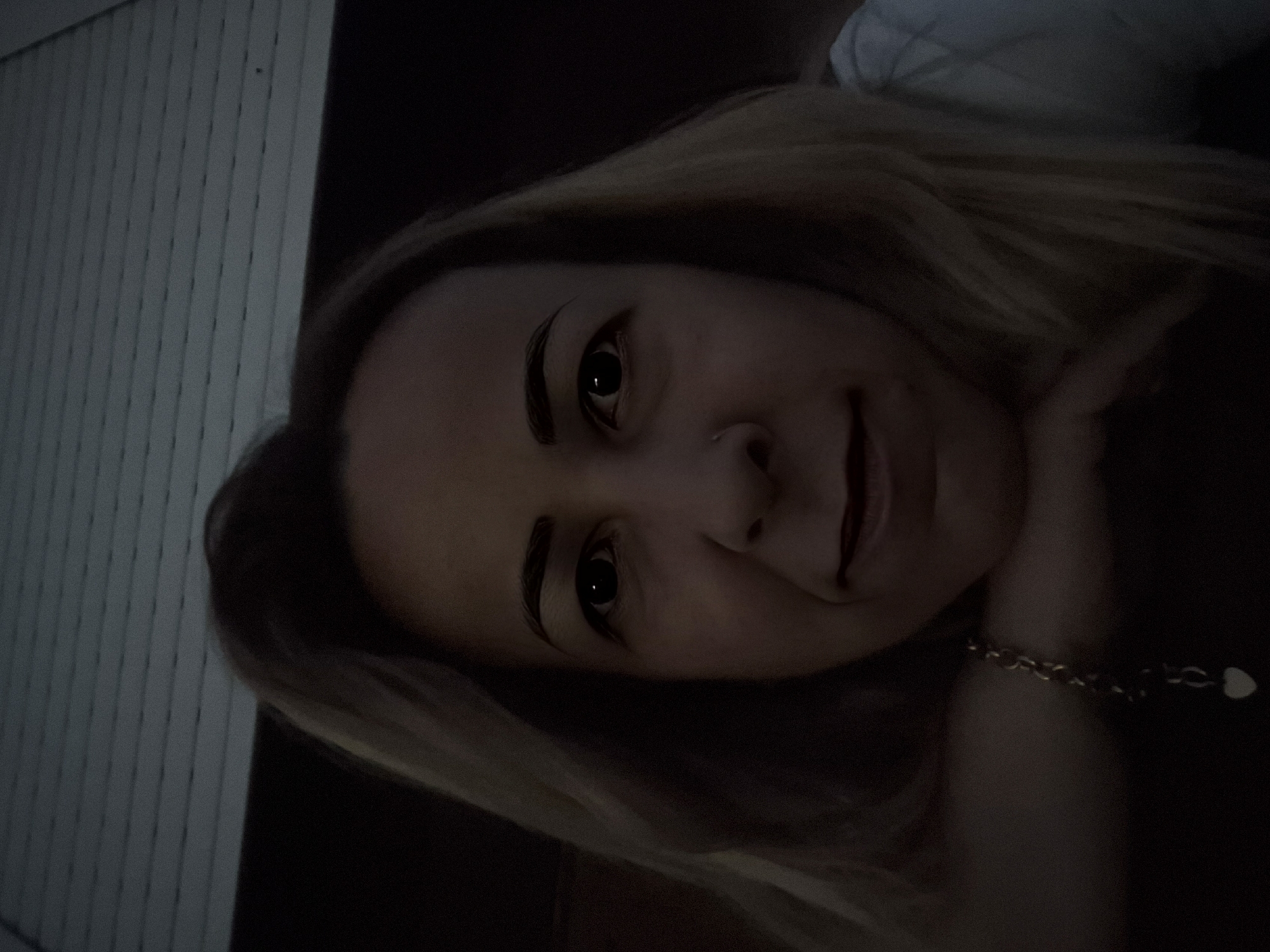 A selfie of a woman with blonde hair is shown. She is leaning her chin on her hands, and her hair falls around her shoulders. She is wearing a gold chain with a heart pendant. The background is dark, but it appears to be outdoors, possibly on a porch or patio, with a closed blind in the upper portion of the image. The lighting is dim, casting shadows on her face.