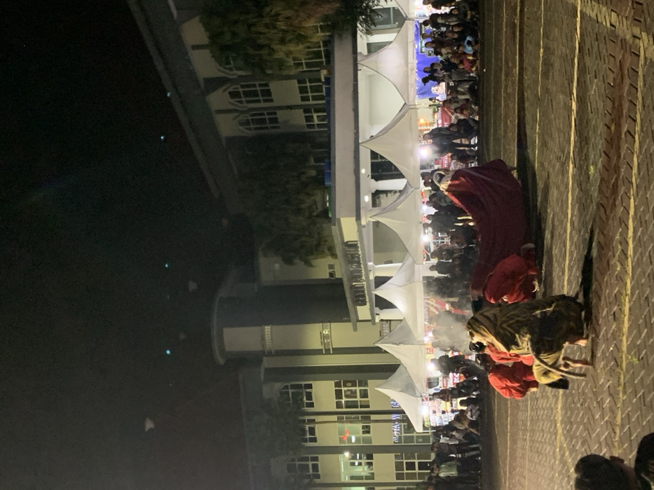 A night performance takes place in front of a large building in Kecamatan Batu, Indonesia, located at coordinates -7.867, 112.514. Five white tent canopies are set up in front of the building. A person wearing a red and gold lion costume performs a dance in the street, their tail dragging behind them. Other performers are wearing red clothing. Spectators are seated on the ground, watching the performance. There are people in the background standing and sitting, some holding drinks. The building is lit with bright lights, and the street is paved with brick. There is smoke coming from the ground near the performers.