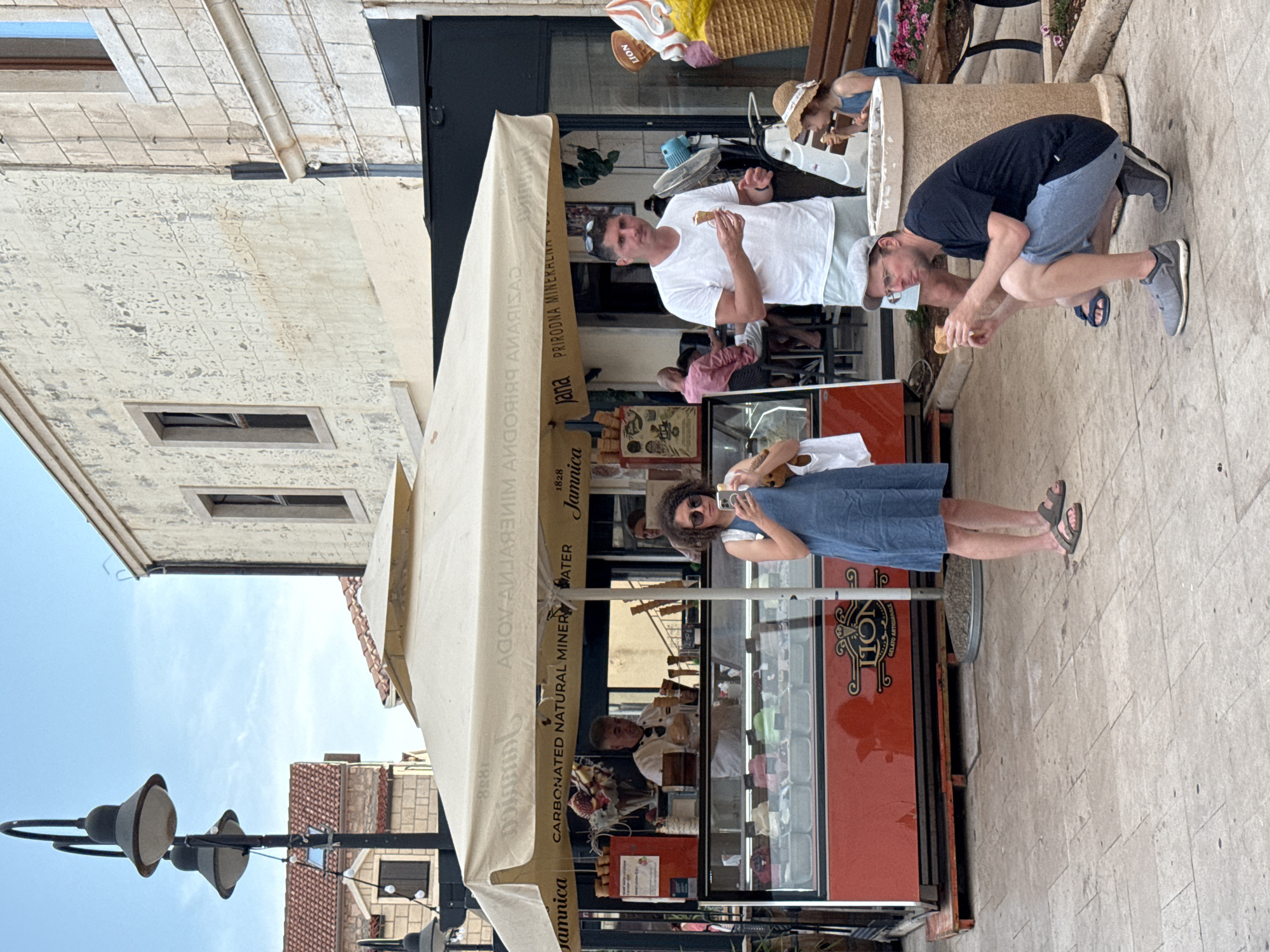 In Primošten, Croatia, an outdoor ice cream stand operates under a large, beige awning. The awning is printed with text, including "Jamnica" and "CARBONATED NATURAL MINERAL WATER". A man in a white shirt and light shorts stands facing forward, holding an ice cream cone. To his right, a young girl in a straw hat and blue dress sits on a bench, eating an ice cream cone. In the foreground, a man in a dark t-shirt and light blue shorts squats down, also eating an ice cream cone. To the left, a woman in a blue dress is looking at her phone and holding an ice cream cone. Behind the awning, an ice cream display case is visible, with various flavors of gelato. A man in a white uniform stands behind the counter, appearing to serve customers. The scene is set on a paved outdoor area, with buildings in the background and streetlights visible.