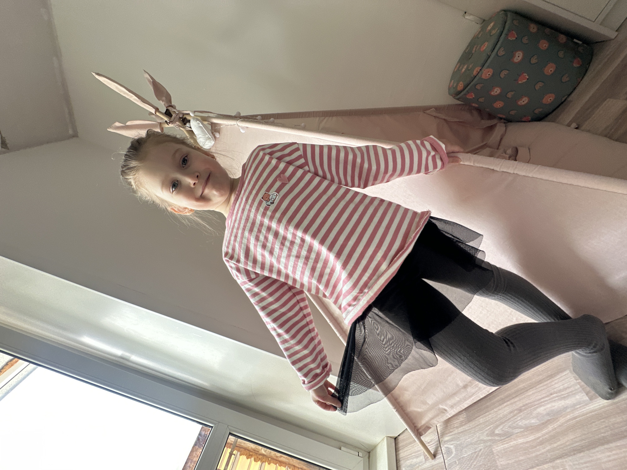 A young girl stands inside a pink teepee. She wears a pink and white striped long-sleeve shirt, a black tulle skirt, and grey ribbed leggings. A printed storage cube is visible behind the teepee. The scene is illuminated by natural light coming from a window. The location is in a building located in the city of Chaikovsky, Russia.