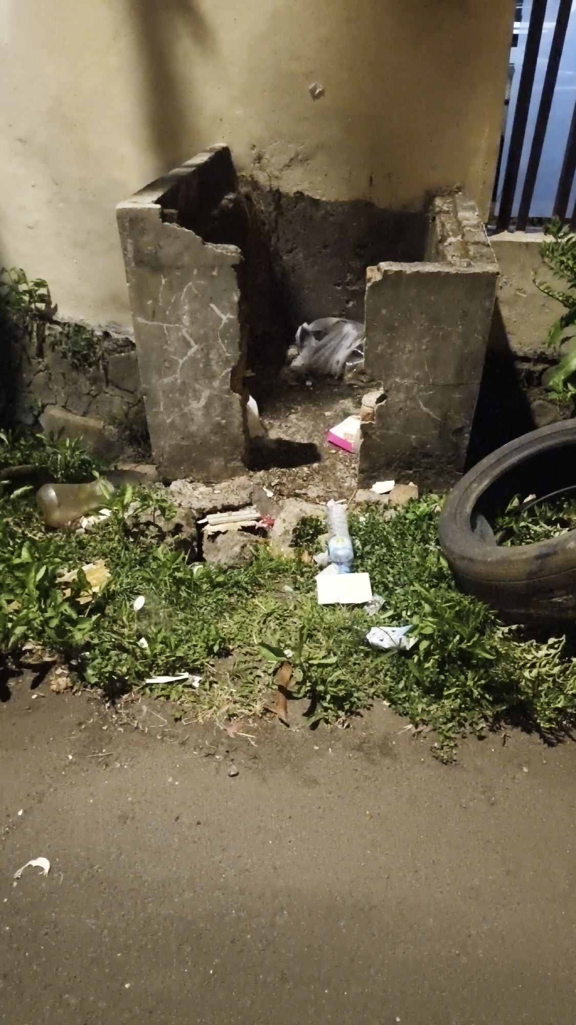 The image depicts an outdoor scene in South Jakarta, Indonesia, focusing on a damaged, concrete structure that appears to be an abandoned or neglected bin or enclosure. The structure is situated next to a light-colored wall and surrounded by overgrown green vegetation. The ground in front of the structure is paved.
The concrete structure itself is fragmented and broken, with some sections missing or eroded. Inside the enclosure, there is a crumpled plastic bag and some other debris. Scattered around the base of the structure and in the surrounding grass are various items of litter, including plastic bottles, paper scraps, and what appears to be a deflated tire to the right of the frame.
The lighting suggests it is nighttime or dusk, with artificial light sources casting shadows. The overall impression is one of disrepair and neglect, with a significant amount of trash accumulating in the area. There are no people visible in the image. The environment appears to be a public space that has fallen into disuse or is not being properly maintained.