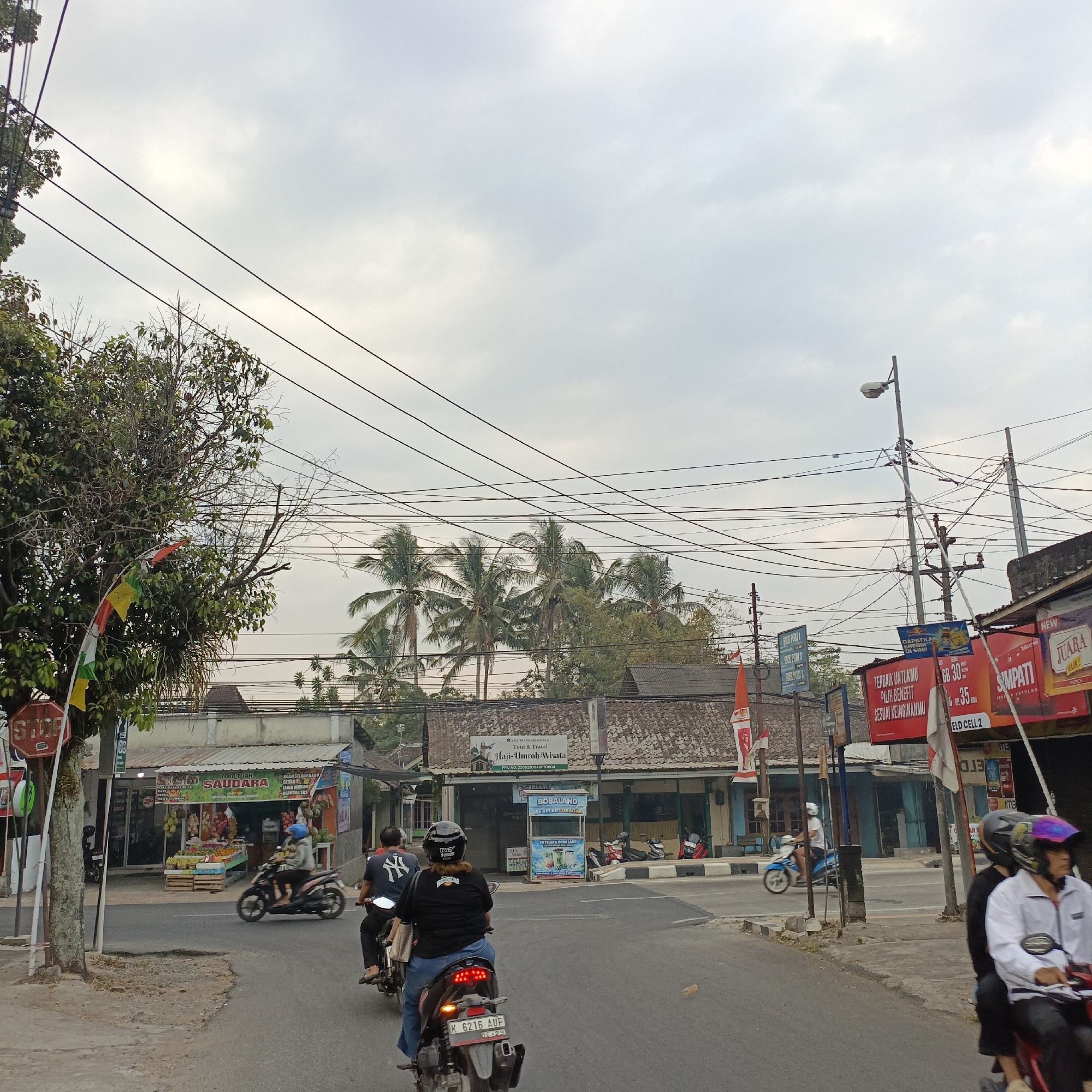 This image captures a street scene in Salatiga, Indonesia. The perspective is from a moving vehicle, looking forward and slightly to the right. The road is paved and appears to be a two-lane street.
In the foreground, two motorcycles are visible, both carrying two passengers. The motorcycle on the left has a rider wearing a black shirt with a white Yankees logo and a black helmet, and a passenger behind them wearing a black shirt and a helmet. The motorcycle on the right has a rider wearing a black shirt and a helmet, and a passenger behind them wearing a white jacket and a colorful helmet. The rear motorcycle has a license plate reading "K 6216 AUF".
Along the right side of the road, there are buildings with shops. One shop sign reads "TOKO BUAH SAUDARA", indicating a fruit shop. Another sign displays "BOBALAND ES TELURURAGAN". Several advertisements are mounted on poles, including one for "SIMPATI" and another with "JUARA".
On the left side of the road, there's a tree with colorful flags hanging from it and a red "STOP" sign. Further down the street, more buildings are visible, some with tiled roofs, and several palm trees are scattered throughout the landscape. Overhead, a dense network of electrical wires crisscrosses the sky, with a street lamp visible on the right side. The sky is overcast with a pale, diffused light. The overall impression is a typical urban street in Indonesia, with everyday activities like commuting by motorcycle and commercial activity.