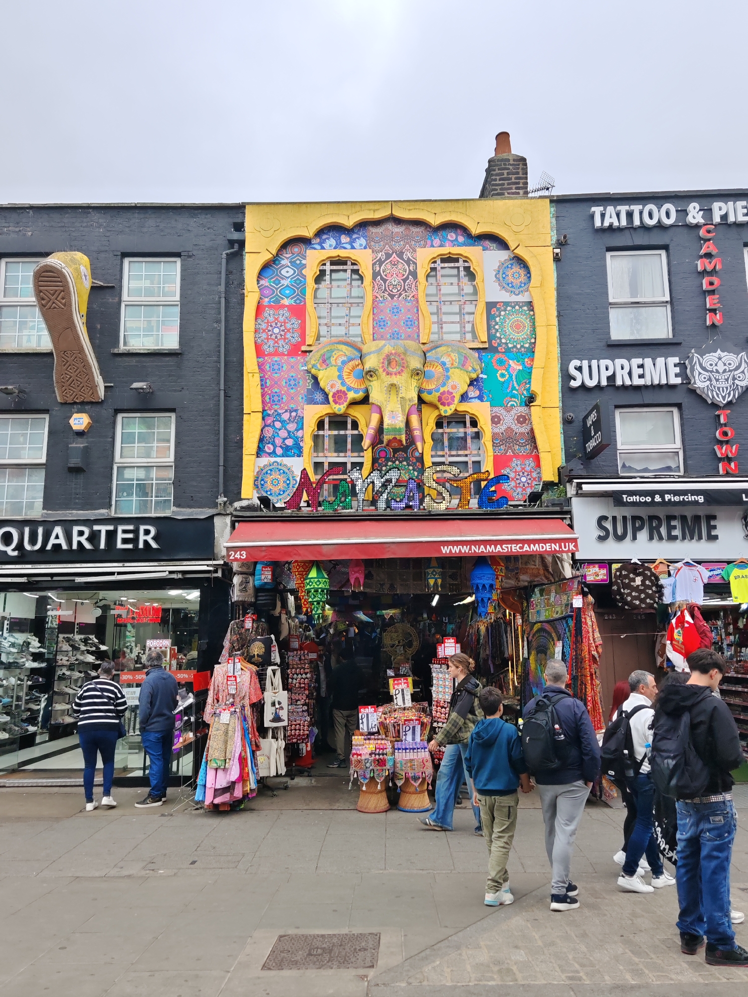 This image captures a vibrant street scene in Camden Town, London, United Kingdom. The architecture is eclectic, featuring a mix of dark brick buildings and brightly decorated facades. A store with a large, ornate elephant head sculpture on its upper facade dominates the view, flanked by a shoe store and a tattoo parlor. People are walking along the street, browsing the shops and merchandise displayed outside. The storefronts are densely packed with goods, creating a bustling atmosphere. The sky is overcast, casting a soft light over the scene.