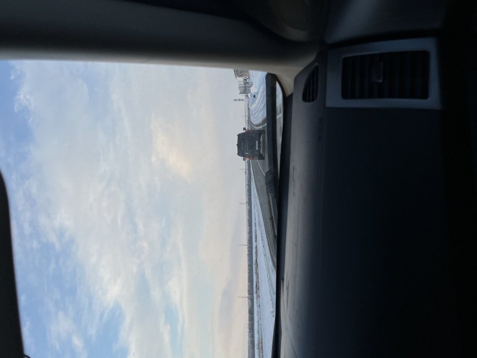 The scene depicts a winter road view from inside a moving vehicle in Anapskaya, Russia. The lower half of the frame shows the dark dashboard and the base of a windshield wiper, indicating the observer's position within a car. Ahead, a dark-colored SUV, identifiable by its rear lights and a partially visible Russian license plate, travels on a clear, dark asphalt road. The road is bordered by snow-covered shoulders and flat, snow-covered terrain, stretching into the distance. A faint line of leafless trees or bushes and utility poles are visible along the horizon. The sky above is partly cloudy, featuring a mix of white and light blue hues. No people are explicitly visible.