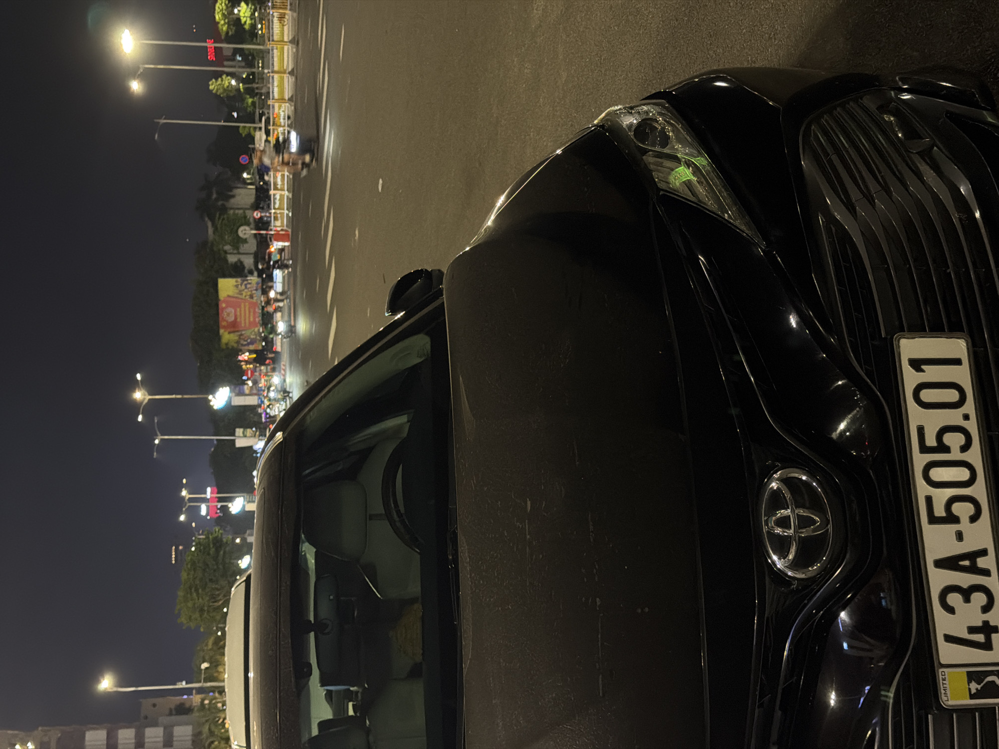 A black Toyota Camry is parked at night on a city street in Da Nang, Vietnam. The car is viewed from the front, with its hood and the driver's side front fender in the foreground. The car's license plate, which reads "43A-505.01," is visible. The interior of the car, including the steering wheel and seats, is also visible. In the background, there are streetlights, traffic, and buildings, suggesting a busy urban environment. The street is wet, indicating recent rain.