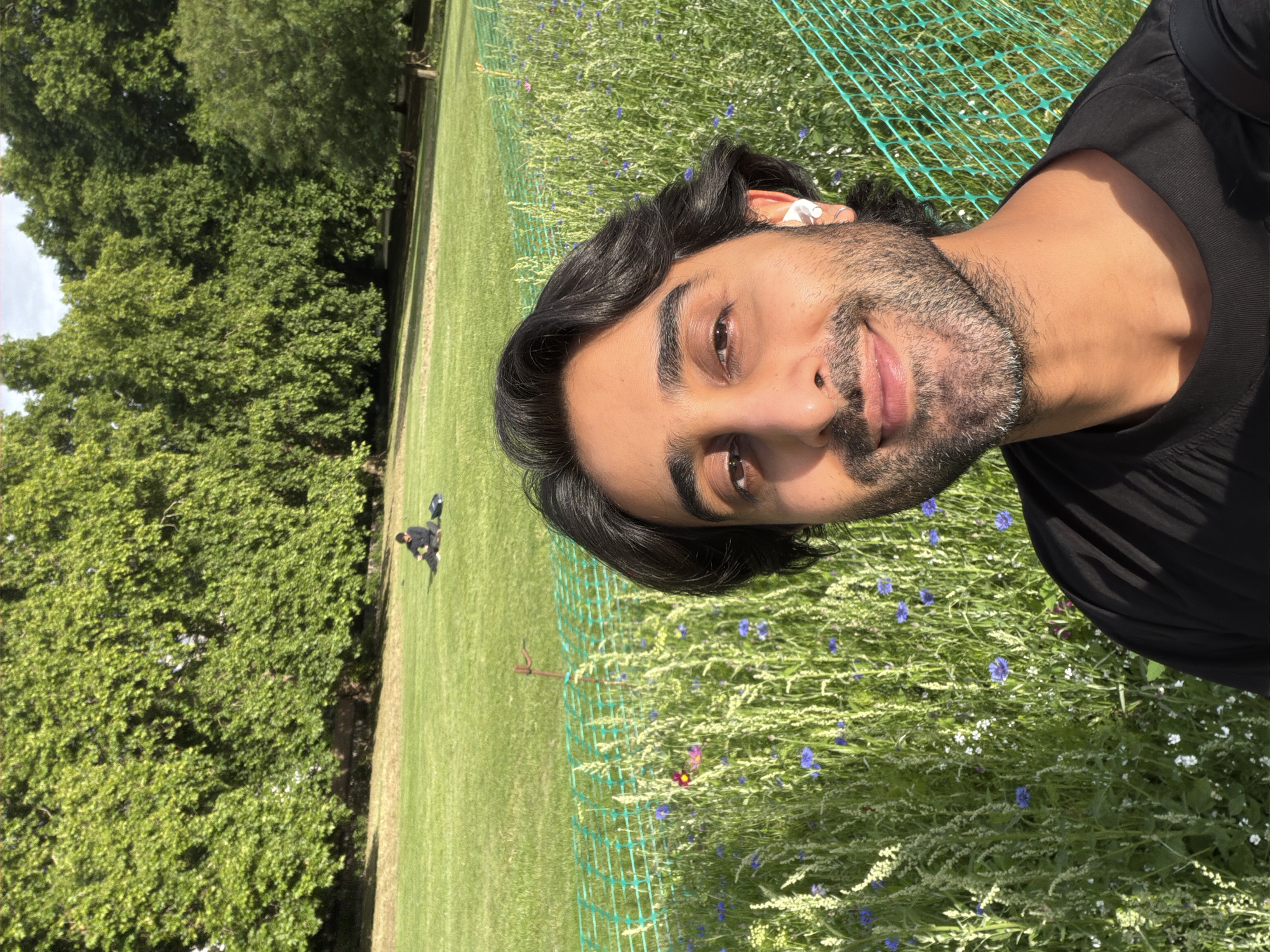The image captures a close-up selfie of a man with dark hair and a beard, wearing a black t-shirt and white earbuds. He is smiling and looking towards the camera. Behind him, a vibrant field of wildflowers, featuring blue and white blossoms, is visible, separated by a green mesh fence. In the background, a lush green lawn stretches towards a line of mature trees, and a person is seated on the grass in the distance. The lighting suggests it is daytime, with sunlight illuminating the scene. The presence of manicured lawns and established trees indicates the location is likely a park within London.