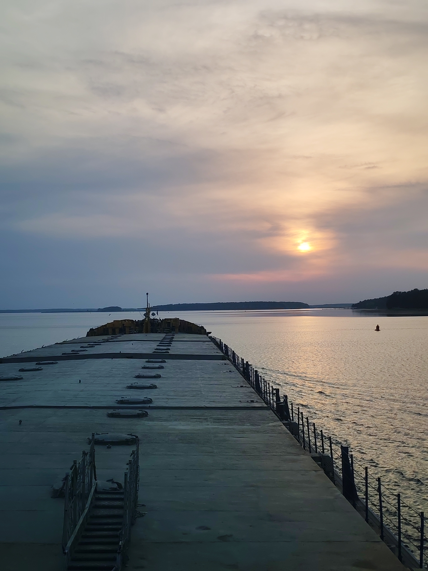 A long, flat concrete structure stretches into a body of water. The structure has what appears to be access hatches along its surface and a metal railing along the right side. To the left, stairs lead up to the surface. In the background, land is visible across the water. An orange buoy floats in the water to the right. The sky is overcast with the sun visible through the clouds. The time of day appears to be dusk or dawn. The location is a pier or dock extending into a large body of water in an unidentifiable city.