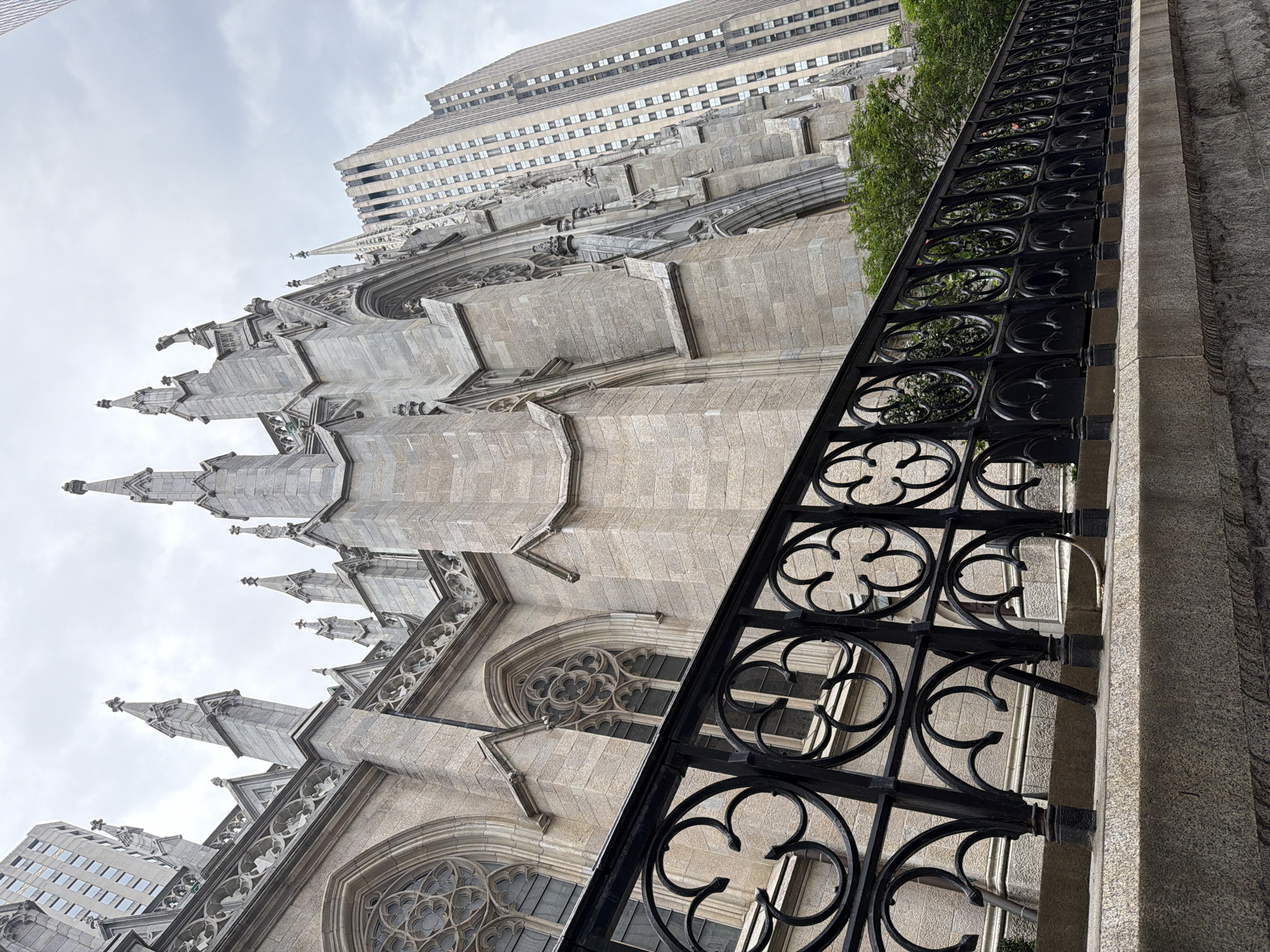 The photograph depicts a ground-level, low-angle view of St. Patrick's Cathedral in New York City, with a foreground focus on a decorative black metal fence. The cathedral, constructed from light gray stone, features gothic-style architecture with spires, pointed arches, and ornate window details. A modern high-rise building is visible in the background, contrasting with the cathedral's historic aesthetic. The sky is overcast, providing soft, diffused lighting. The environment appears to be a street or walkway adjacent to the cathedral, with the metal fence acting as a boundary or barrier.
