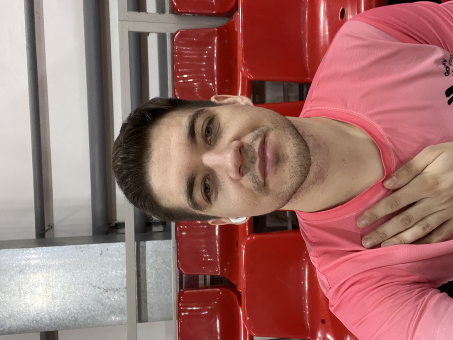 A selfie captures a man in a pink athletic shirt, seated in a red stadium seat. He has short, dark hair, light skin, and a trimmed beard. An earpiece is visible in his ear. His hand rests on his chest. Rows of empty red seats fill the background, alongside gray metal railings and a white wall, suggesting an indoor stadium setting. The image appears to be slightly rotated.