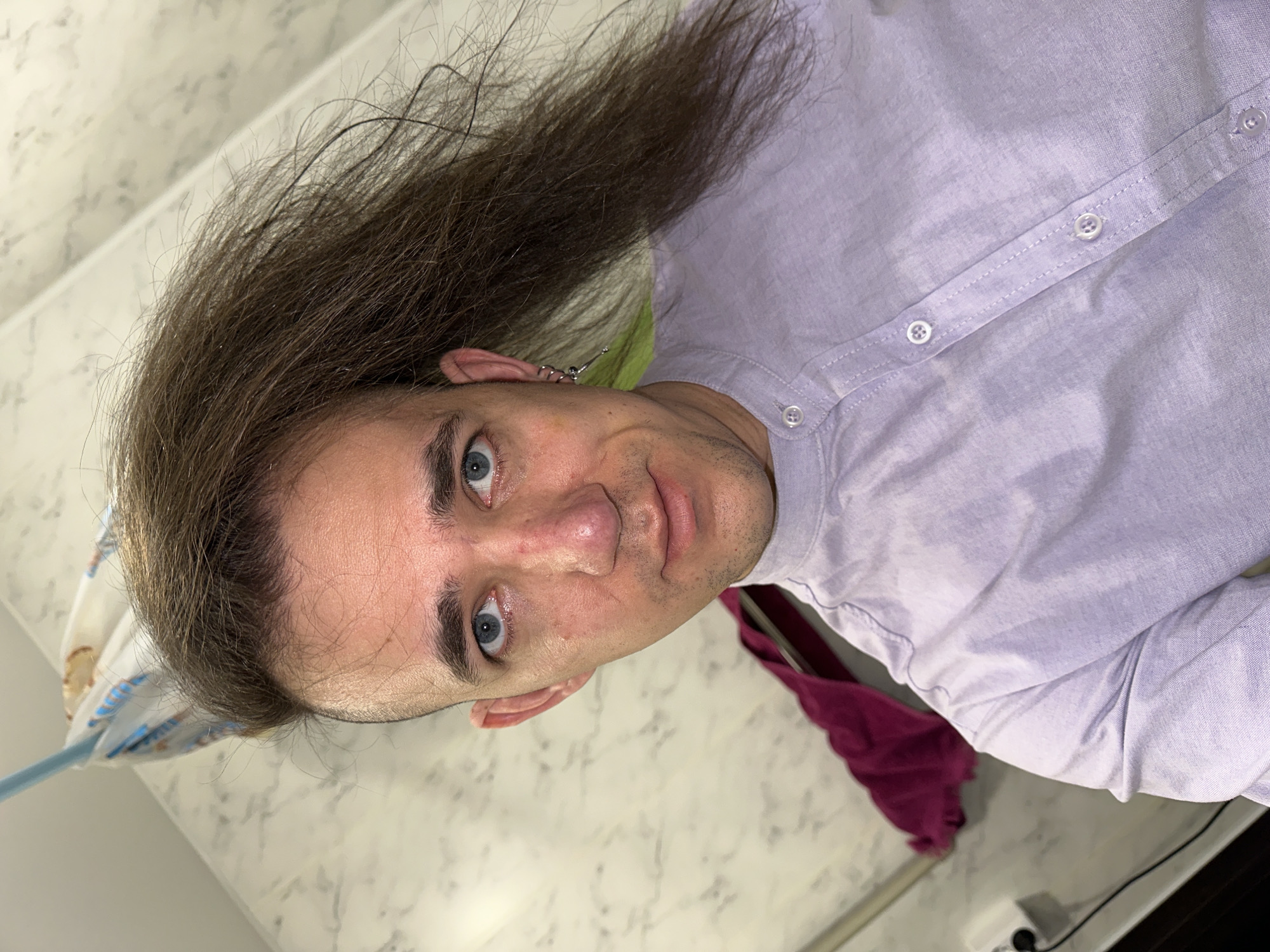A selfie taken by a male with blue eyes and brown hair, wearing a light purple button-down shirt. The background shows a light-colored countertop and a blurred object in the lower right corner. There is no indication of Saratov, Russia, or any other specific location in the image.