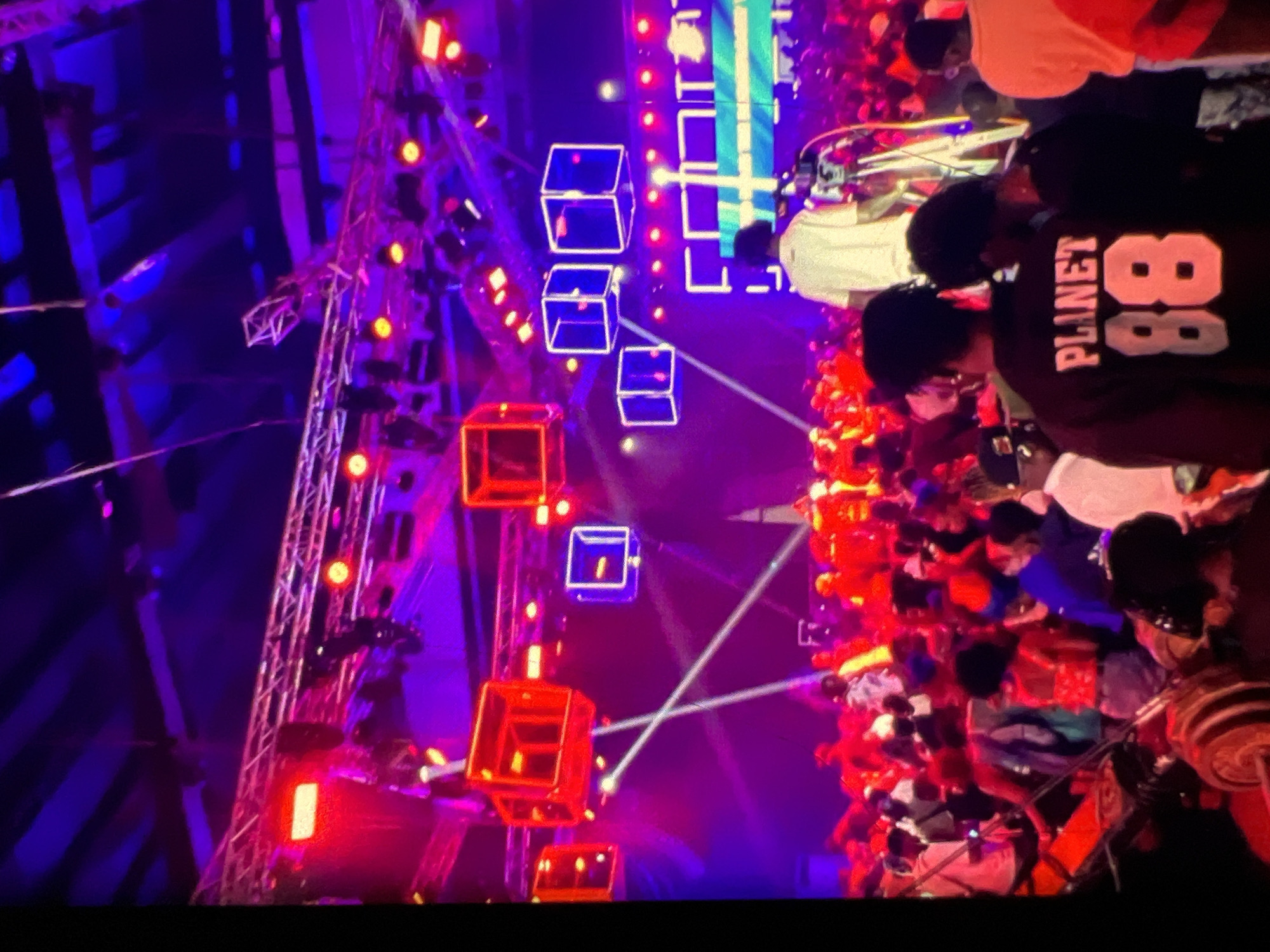 The image depicts an indoor event, possibly a concert or festival, illuminated by vibrant red and purple lights. Several large, illuminated cubic structures hang from the ceiling, some with red lighting and others outlined in blue. Stage lighting and truss systems are visible. In the foreground and midground, a large crowd of people are gathered. Many are dressed in casual attire, and some are wearing distinctive clothing, such as a person in the foreground wearing a dark sweatshirt with "PLANET 88" emblazoned on the back. There appears to be a stage area with equipment like cameras and microphones. The overall atmosphere suggests a lively and energetic event. This scene is consistent with various entertainment venues in Lagos, Nigeria, known for hosting large-scale music and cultural events.