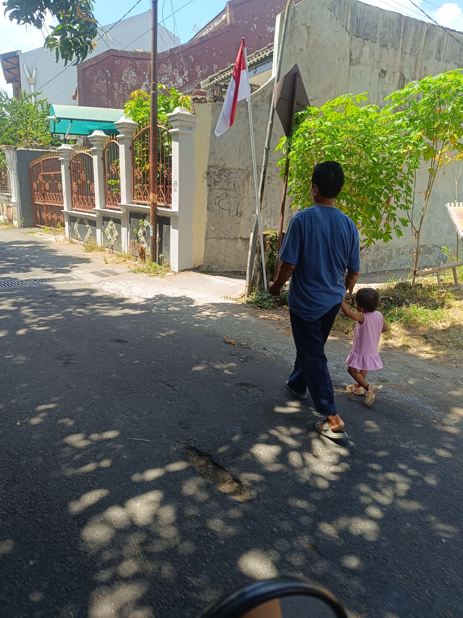 In Yogyakarta, Indonesia, a man in a blue t-shirt and dark pants walks down a street, holding the hand of a small child in a pink dress. The man is wearing sandals, and the child is wearing light-colored sandals. They are walking away from the camera on a paved road with shadows cast by overhead trees. Behind them, a fence with ornate metalwork borders a property with a green awning and trees. A pole with a red and white flag, likely the Indonesian flag, is visible to the left of the man. The background includes a textured wall and more greenery.