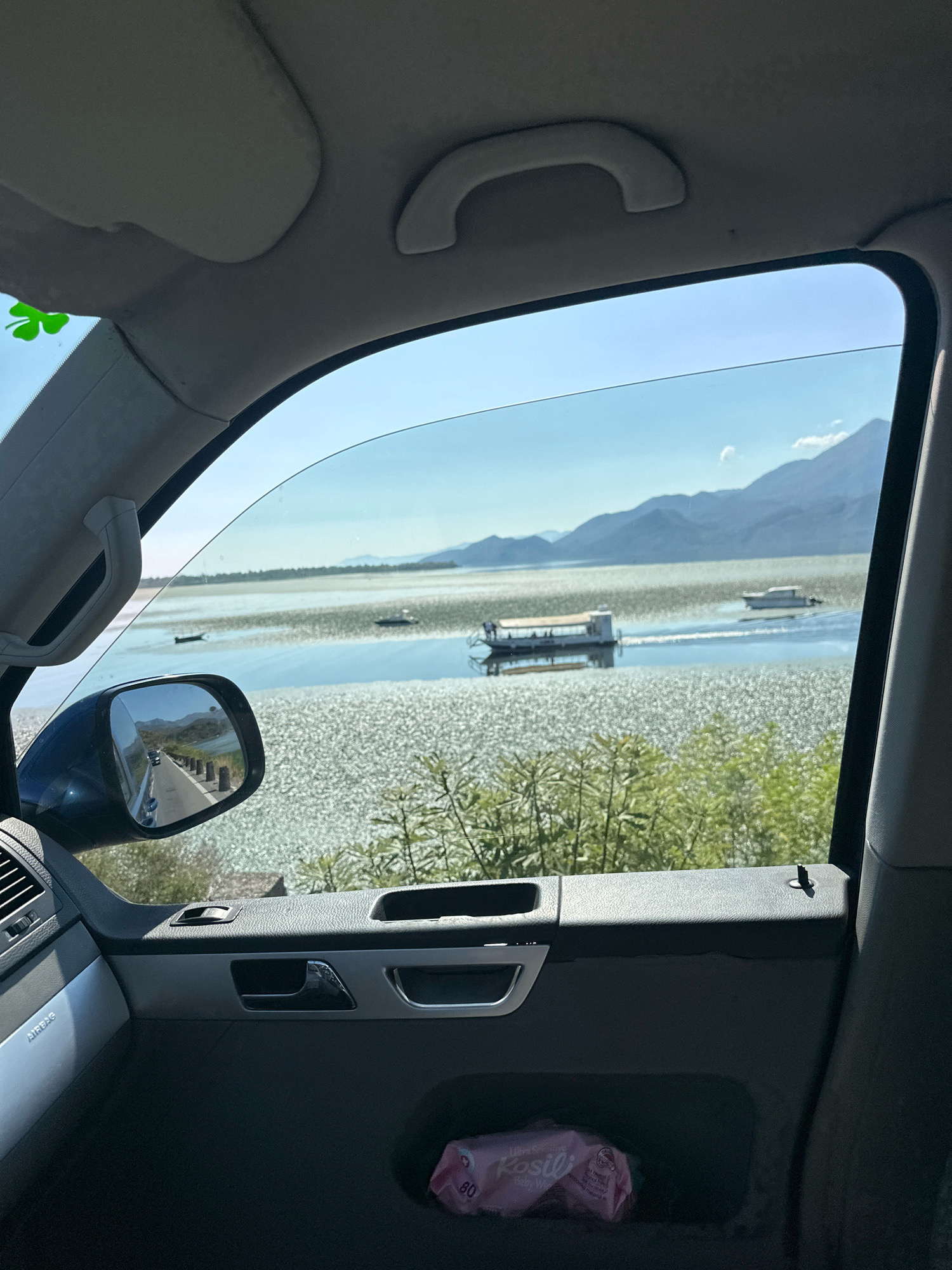 A wide shot taken from inside a vehicle in Vranjina, Montenegro, shows a scenic view of a lake with several boats and mountains in the background. The foreground shows the interior of the car, with the dashboard, door panel, and a portion of the car's ceiling visible. A green shamrock sticker is affixed to the windshield near the top left corner. The side mirror reflects a road with passing cars and a tree-lined embankment. In the middle ground, two boats are on the water, one appears to be a passenger boat with people on board, and another smaller boat is further out. The lake is covered with a dense layer of aquatic vegetation that reflects the sunlight, creating a shimmering effect. In the background, a range of mountains under a clear blue sky complete the landscape. A packet of baby wipes is visible in the door pocket.
