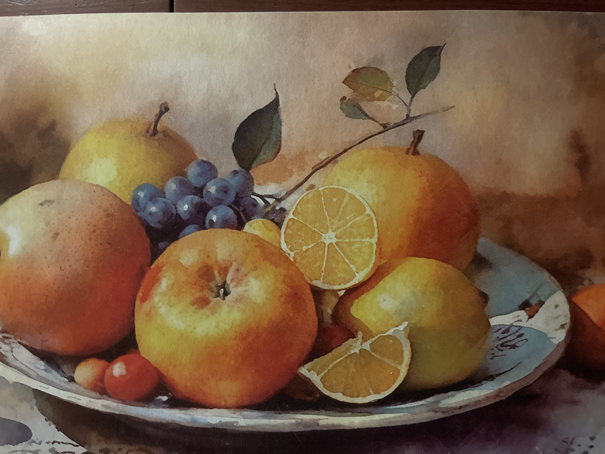 This is a still life painting of a bowl of fruit. The fruit includes apples, pears, grapes, oranges, and possibly lemons. The fruits are arranged in a silver bowl. The painting has a soft, warm color palette with oranges, yellows, and blues predominating. There are also some green leaves and stems visible. The background is a soft gradient of beige and brown, suggesting a diffused light source. The painting does not depict any people, actions, or interactions. It is a static composition focused solely on the arrangement of fruit. There is no information within the image to suggest a location in Minsk, Belarus or any other specific city.