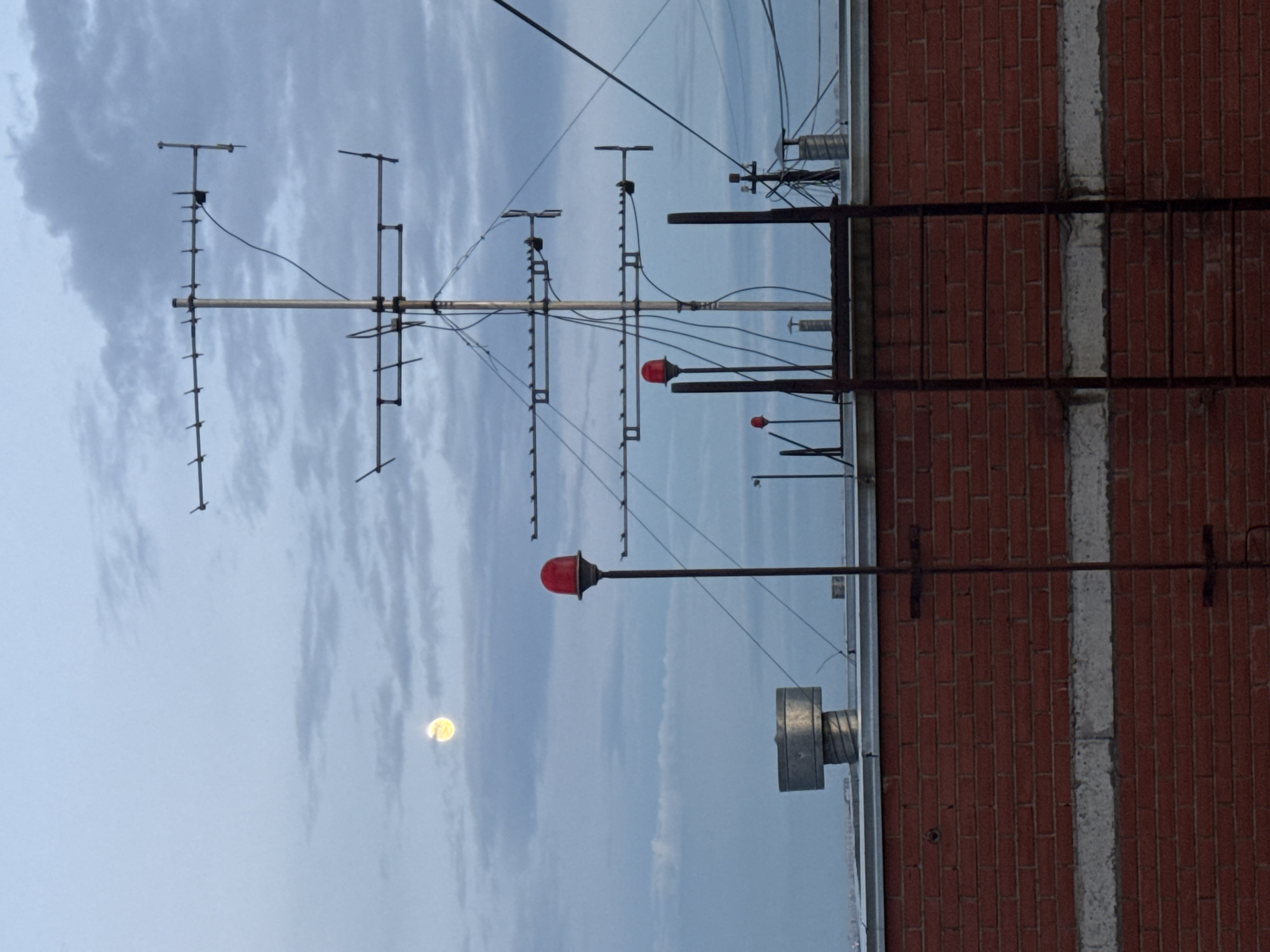 The image captures a rooftop scene with multiple antennas and a brick wall in the foreground, set against a cloudy twilight sky where a partially obscured moon is visible. The location is identified as Khimki, Russia. Prominently featured is a large television antenna with horizontal and vertical elements. To the left of this antenna, a tall pole supports a red flashing light, and to its right, another similar pole also has a red light. A metallic ventilation pipe is visible on the rooftop to the left. A rusty metal ladder is attached to the brick wall, suggesting access to the rooftop. The brick wall is a dominant element in the foreground, with a horizontal concrete band running across it. The sky is overcast with soft, diffused light, typical of early evening or dawn.