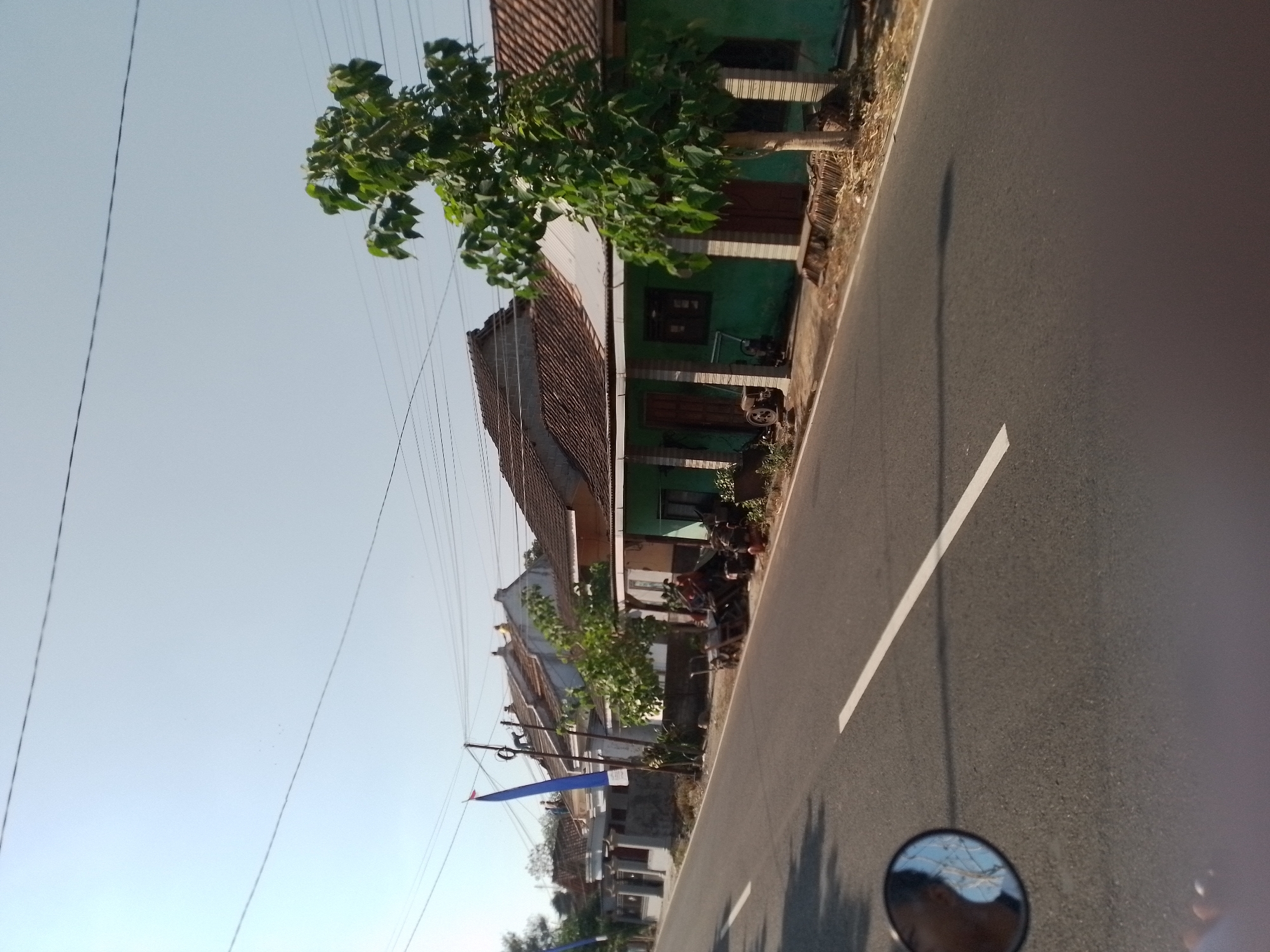 The image depicts a street scene in Ngancar, Indonesia. A paved road with a white line marking its center runs diagonally across the frame from the lower left to the upper right. Along the right side of the road, a row of houses with tiled roofs and green walls is visible. A large tree with dense foliage stands in front of one of the houses. In front of the houses, several agricultural machines, possibly for farming, are parked. A blue flag is attached to a pole in front of one of the houses. The sky is clear and blue. In the lower left corner of the image, a motorcycle's rearview mirror reflects the head of a person and the sky. The overall impression is a sunny day in a rural or semi-rural Indonesian setting.