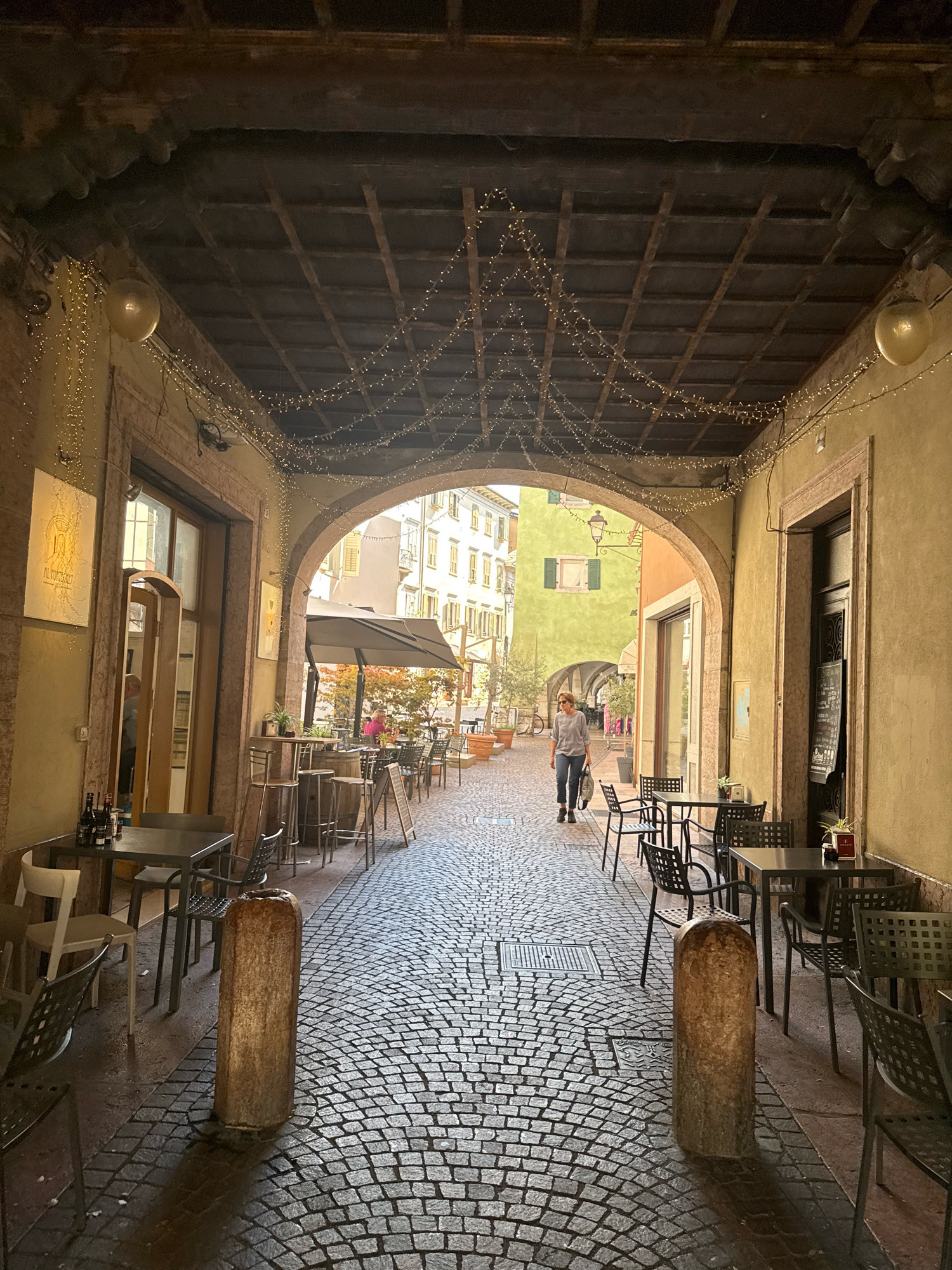 The image captures a view down a covered walkway or arcade in Trento, Italy. The walkway is paved with cobblestones, and the ceiling is made of wooden beams with a grid pattern. String lights are draped across the ceiling, and two large, translucent balloons are visible hanging from above.
On either side of the walkway are buildings with arched entrances and windows. The left side features a restaurant or cafe with tables and chairs set up both inside and outside. One table has bottles of wine on it. The right side also has tables and chairs set out, with a chalkboard menu displayed next to a doorway.
Further down the walkway, a woman in a gray top and dark pants is walking away from the camera. She is carrying a tote bag. Beyond her, the walkway opens up into a street with more buildings and outdoor seating areas. The architecture suggests a historical European city. The overall impression is of a charming, pedestrian-friendly area with outdoor dining.