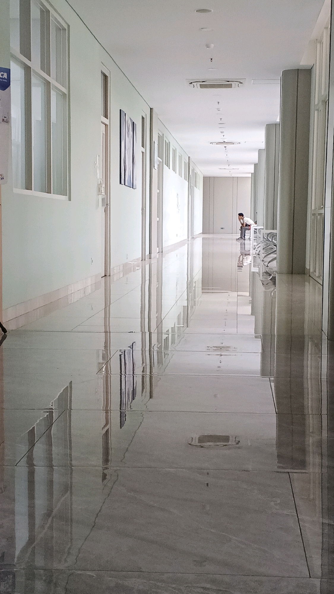 The image depicts a long, well-lit hallway with a highly reflective tiled floor, suggesting it may be a modern building, possibly an office or institutional setting. The hallway has large windows on the left and a series of doors and pillars on the right. The ceiling features fluorescent lighting and air conditioning units. In the distance, a lone individual is seated on a bench, appearing to be looking forward. The reflective floor creates a strong sense of depth and symmetry, with distorted reflections of the ceiling lights and structural elements. The overall impression is one of cleanliness and order. While no specific Semarang landmarks are visible, the architectural style and building materials are consistent with contemporary construction found in urban areas of Indonesia.