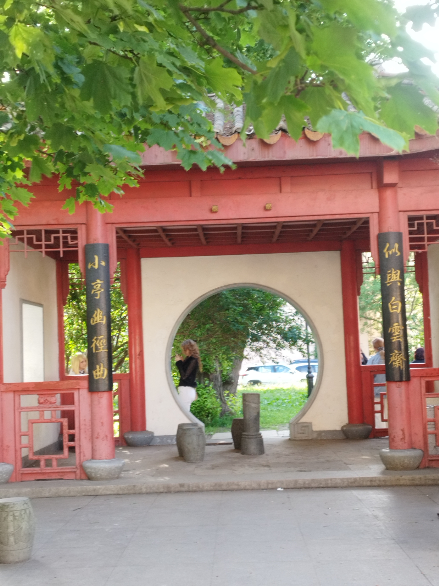 The image shows a portion of a Chinese-style pavilion in a park, likely in Saint Petersburg, Russia, given the context provided by the user's prompt. The pavilion is predominantly red with ornate detailing and is partially shaded by lush green trees. A circular opening in the pavilion reveals a view of greenery and a parked car. Two vertical black signs with gold Chinese characters are attached to the red pillars. On the left side, a woman with blonde hair is visible through a latticed opening. On the right side, a woman with dark curly hair and wearing a black top and white pants is standing in front of the circular opening, facing away from the camera. Several stone decorative elements, resembling barrels or columns, are placed around the pavilion's base. The ground is paved with gray tiles.