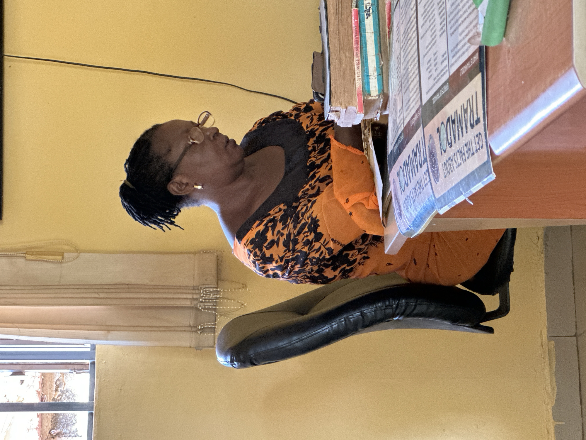 The image shows a Black woman wearing glasses and an orange and black patterned dress, sitting at a desk. She has her hair pulled back in a ponytail. She is looking down at papers on the desk. In front of her is a stack of books. The room appears to be an office, with a yellow wall and a window with a curtain. The desk is made of wood. There are some papers spread out on the desk, with the words "GET THE FACTS ABOUT TRAMADOL" visible on one of them. The image was taken indoors, likely in an office setting in Kaduna, Nigeria, given the context provided.