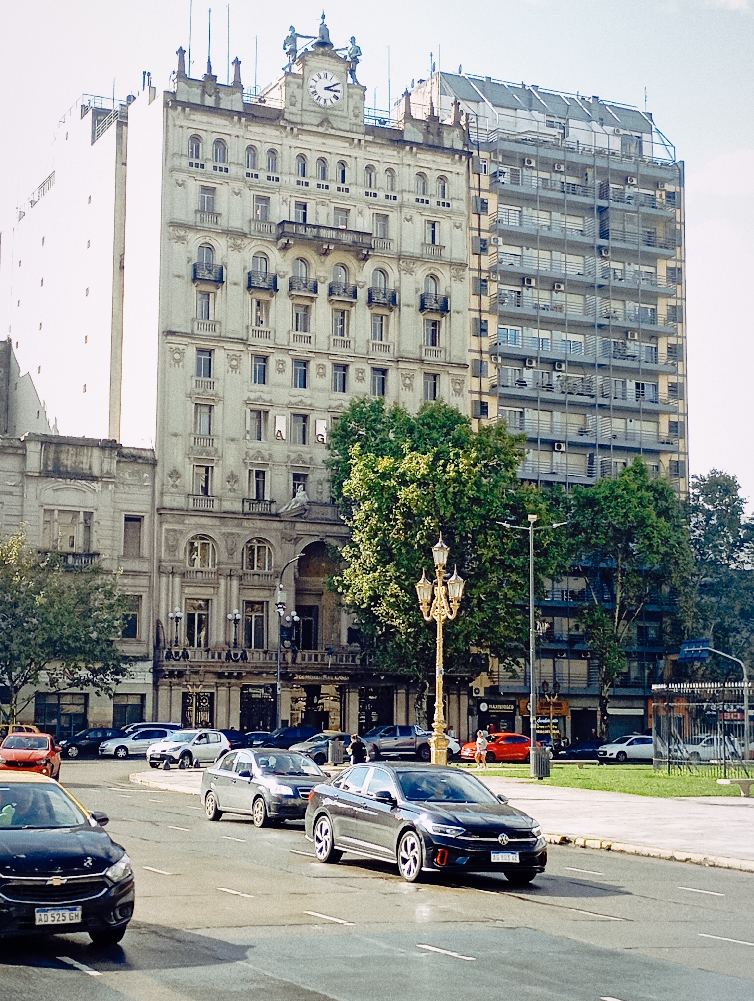 A daytime street scene unfolds in Buenos Aires, Argentina, featuring a mix of architectural styles and active urban life. The dominant structure is a grand, multi-story classical building with ornate detailing, balconies, and a prominent clock tower at its apex. Two statues, possibly representing soldiers or guards, flank the clock face, which shows the time as approximately 11:15 or 11:20. To the right of this classical building stands a contrasting modern, multi-story building characterized by numerous balconies and visible air conditioning units. Further left, a tall, plain white building with small windows is also visible, along with an older, lower-rise building.
The foreground is occupied by a busy multi-lane street with several cars in motion and parked. A dark-colored Chevrolet car is prominent in the lower left, displaying the license plate "AD 525 GH". Another dark gray Volkswagen, further to the right, bears the license plate "AG 803 AZ". An ornate, golden lamppost stands on the right side of the road, partially obscured by lush green trees that line the street.
In the background, on the sidewalks, a few people can be seen, and more parked cars are visible. Signs for businesses are present, including one that reads "MAXIKIOSCO" and another partially visible as "MODULARARA". Banners or signs hanging from a window of the classical building display the letters "A" and "G". The bright sunlight suggests a clear, sunny day. The scene captures the dynamic atmosphere of a city intersection.