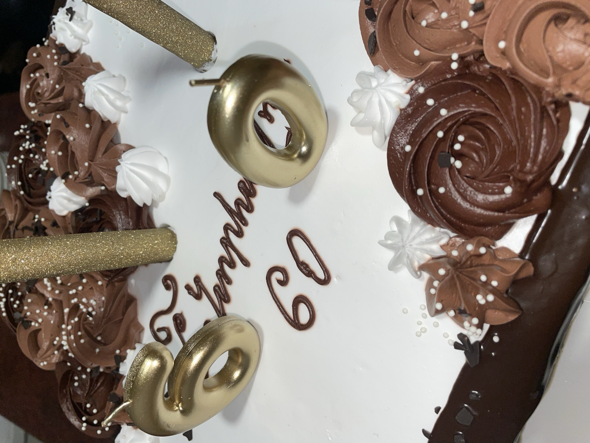 A close-up shot depicts a celebratory cake, likely for a 60th birthday, set against an indeterminate background, but NOT Aratashen, Armenia. The cake features white frosting as its base, adorned with alternating swirls of light and dark chocolate frosting, as well as small white meringue peaks. Edible white pearl sprinkles are scattered across the chocolate frosting. The number "60" is spelled out on the cake, both with gold metallic number candles and with dark chocolate frosting lettering. A message, presumably "Happy Birthday," is also written in chocolate frosting. Two gold glitter candles complete the decoration.