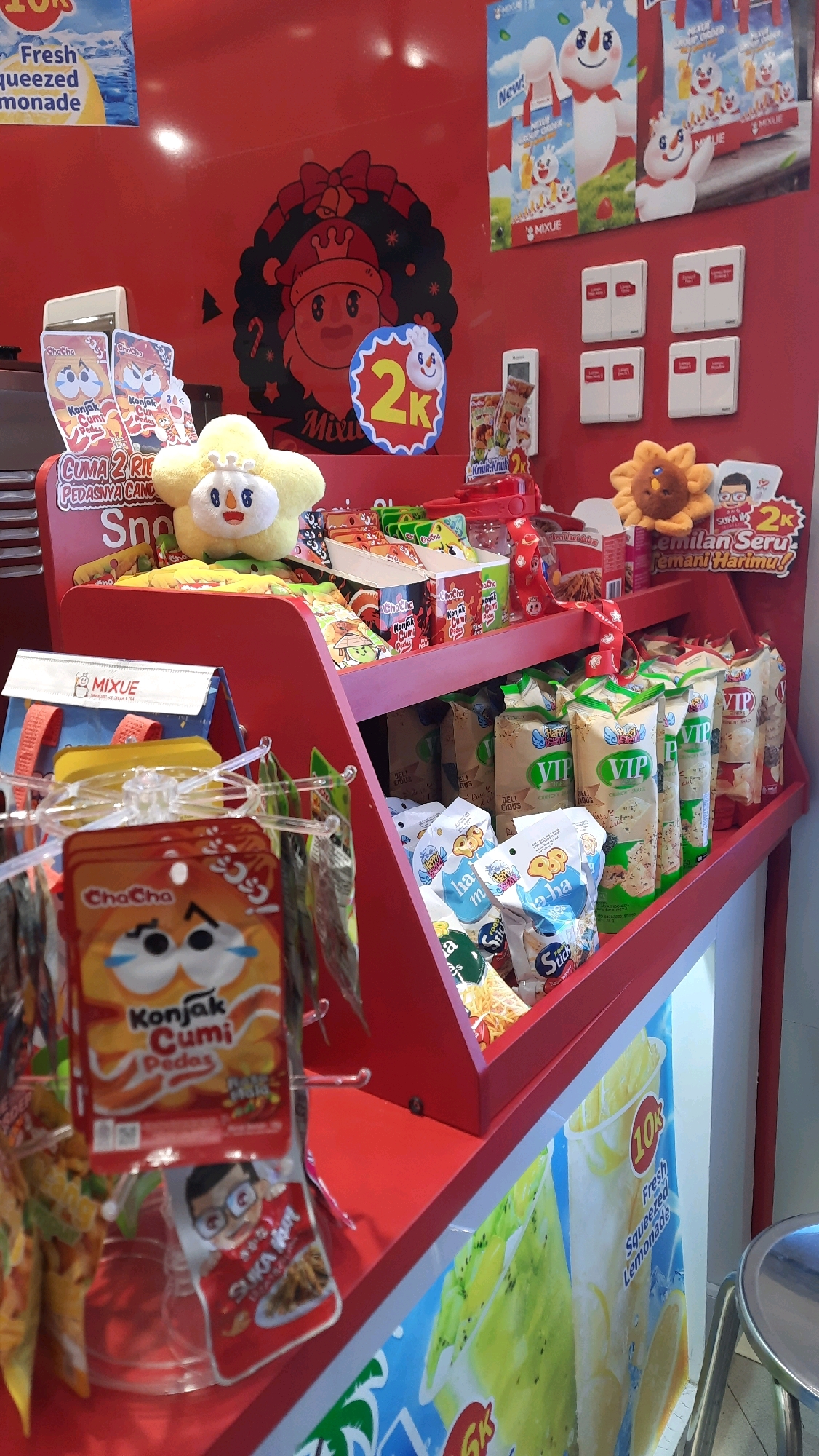 The image shows a red shelf display in what appears to be a retail store in Sidoagung, Indonesia, likely a convenience store or a small market, given the products displayed. The shelves are filled with various snacks and drinks, predominantly from the brand Mixue.
On the top shelf, there are promotional posters featuring Mixue's characters, including a snowman-like figure and a bear. A Christmas-themed decoration is also visible on the red wall behind the display. Several packets of snacks are arranged in cubbies on the top shelf, including "Cha Cha Konjak Cumi Pedas". A yellow plush toy resembling a flower with a smiling face is placed on this shelf.
The middle shelf holds more packaged snacks, with prominent displays of "VIP Crunchy Snack" and other products with colorful packaging. A red toy gun is also visible on this shelf.
The bottom shelf has a selection of smaller snack packets, including "Cha Cha Konjak Cumi Pedas" and other items with cartoonish illustrations. A larger poster featuring iced drinks, specifically "10k Fresh Squeezed Lemonade" and kiwi-flavored drinks, is displayed at the base of the counter.
The lighting suggests an indoor environment, and the overall impression is of a brightly lit point of sale for impulse purchases, characteristic of Asian convenience stores. There are no people visible in the image.
