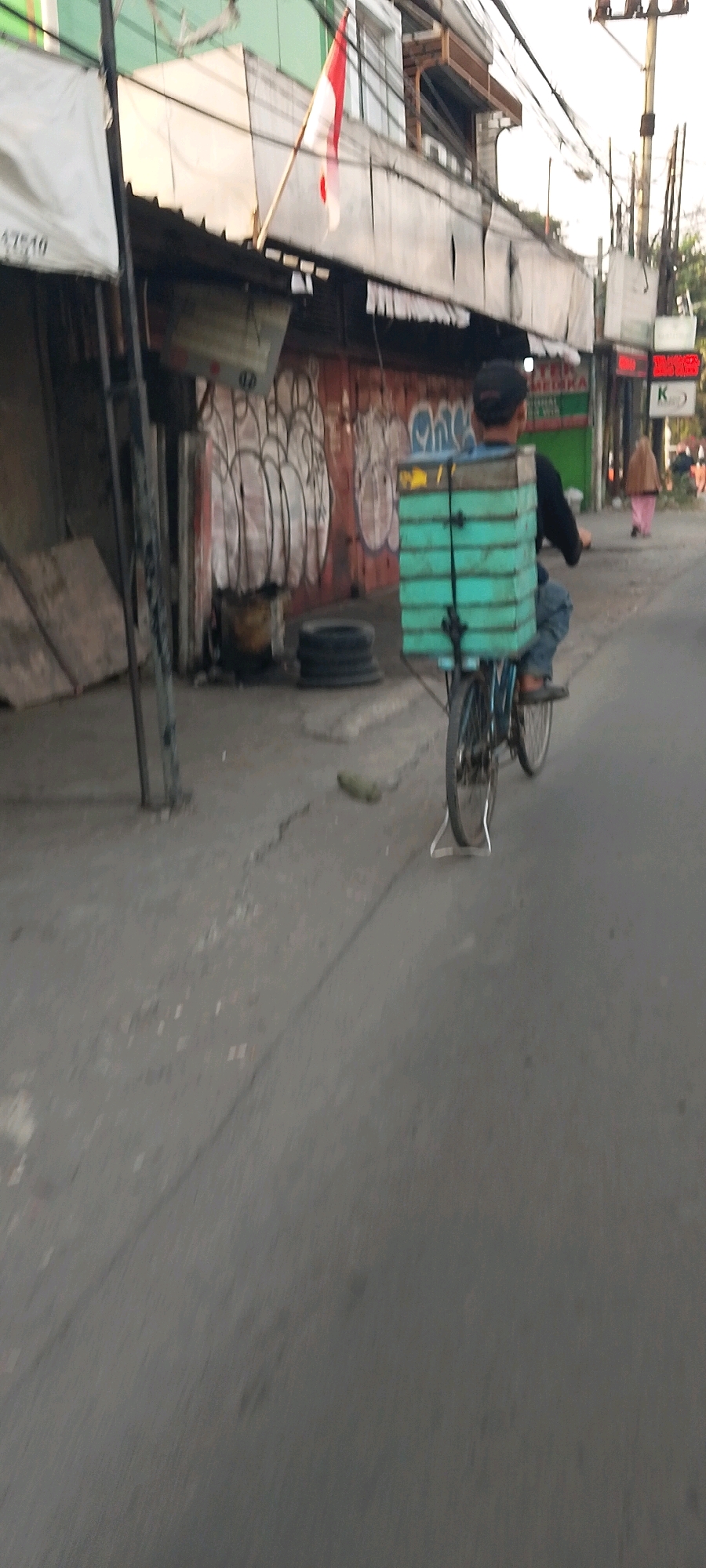 In Karangsatria, Indonesia, a person wearing a dark cap and blue jeans is riding a bicycle on a paved road. The bicycle is loaded with a stack of light blue rectangular containers secured to the rear rack. The person is facing away from the camera and appears to be in motion. Behind the cyclist, a building with a corrugated metal roof is visible, adorned with graffiti. A red and white Indonesian flag is displayed on a flagpole attached to the building. Further down the road, another person in a tan headscarf and pink clothing is walking away from the camera. Overhead, a complex network of electrical wires is strung between poles. The environment suggests an urban or semi-urban setting.