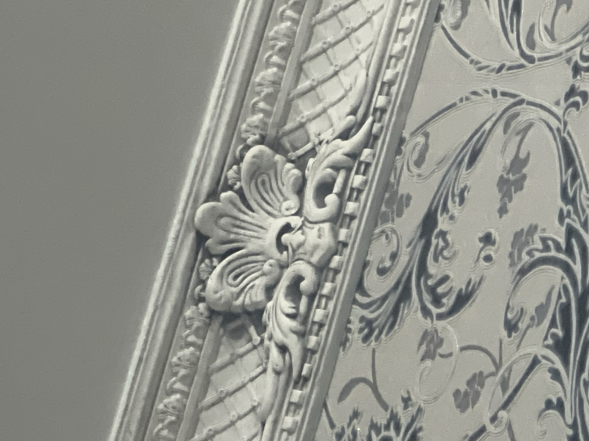 This is a close-up, high-angle, black and white photograph of ornate architectural detailing. In the foreground, a ceiling or upper wall section is visible, adorned with intricate moldings and a repeating floral wallpaper pattern. The molding at the top of the frame features a relief carving of a stylized shell or fan shape flanked by acanthus leaves, resting above a textured band with a diamond-like pattern. Below this, the wallpaper displays a dark blue or grey, swirling floral design against a lighter background. The lighting is soft, casting minimal shadows and highlighting the textures of the relief and the wallpaper. The image is sharp and focused on the decorative elements, with the upper part of the frame showing a plain, out-of-focus gray area that likely represents the ceiling. The photograph does not contain any people, actions, or interactions. The location is identified as Akhalkalaki, Georgia, suggesting this is an interior architectural detail from a building in that city.
