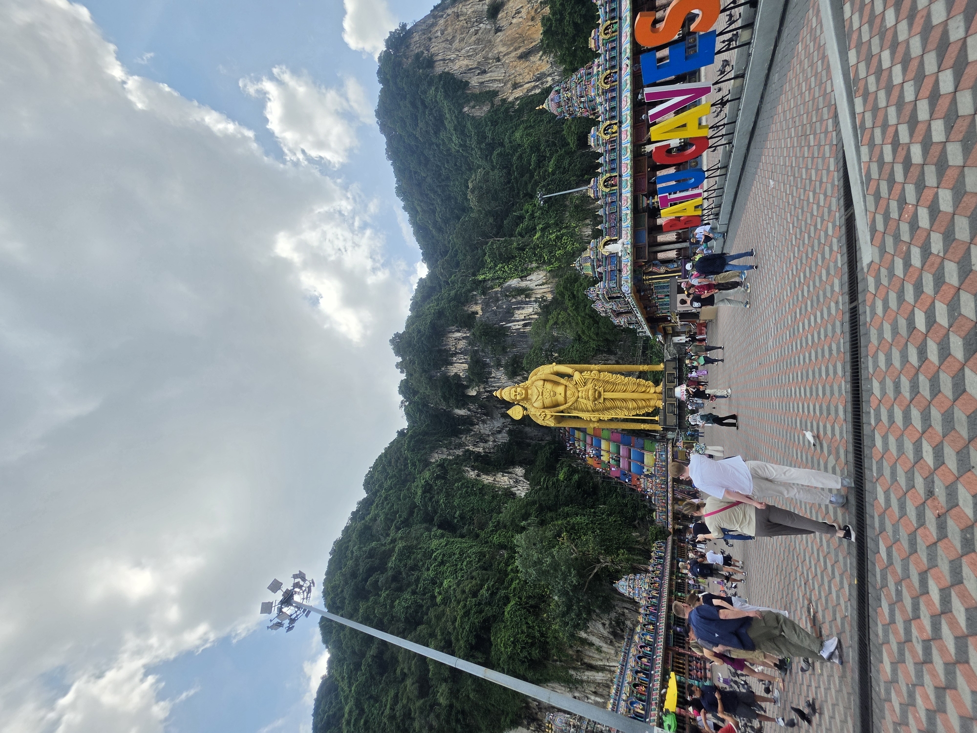 This photograph captures the exterior of the Batu Caves in Selayang Municipal Council, Malaysia. A large, golden statue dominates the foreground, set against a backdrop of a green, rocky mountain. The foreground is populated by a crowd of people walking on a geometrically patterned tiled path. Colorful architectural structures can be seen to the right of the path. The sky above is a mix of blue and cloudy skies. A tall light pole is visible on the left side of the image. A large, colorful sign is to the top right.