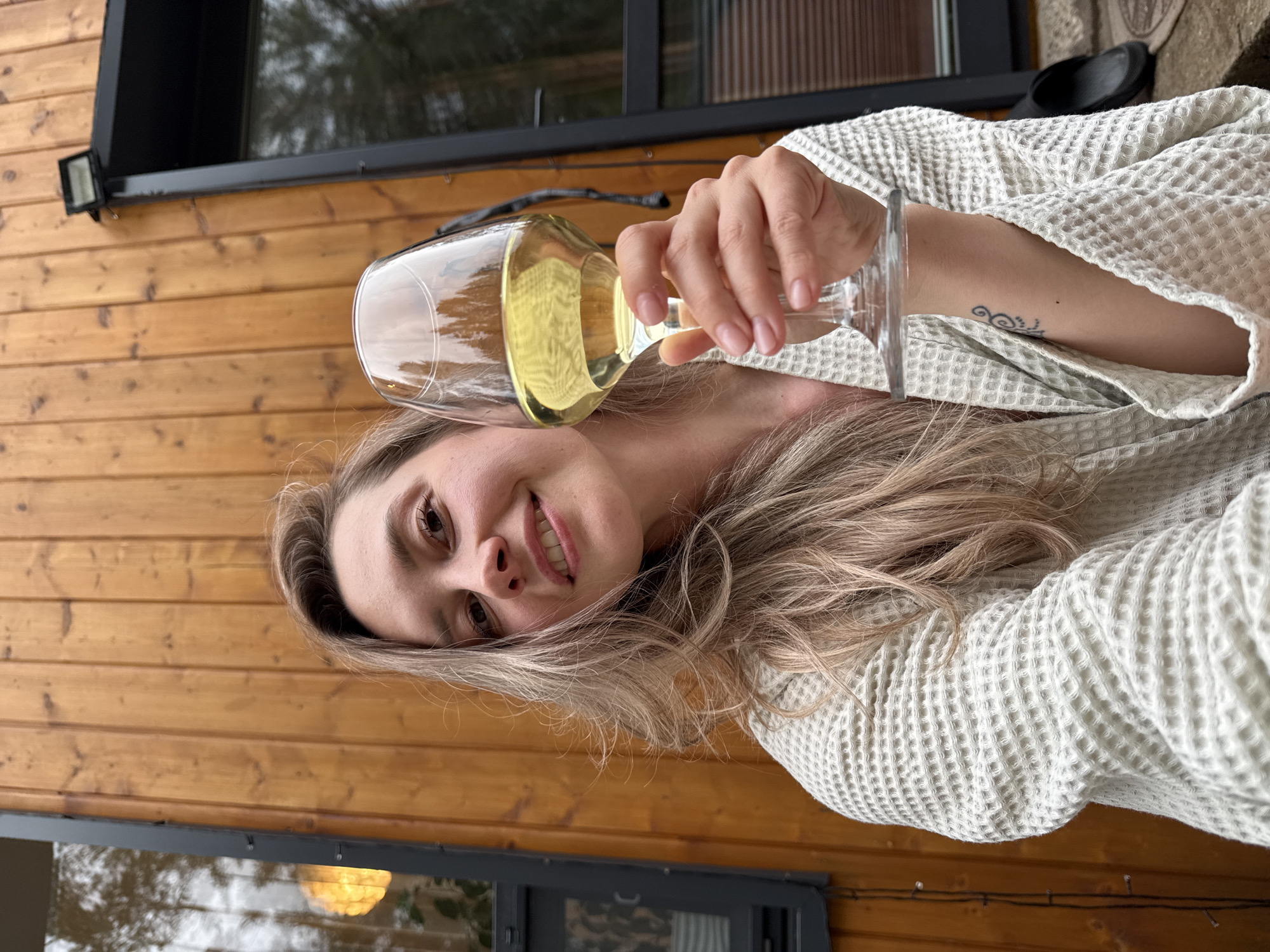 A woman in a white waffle-textured robe is holding a wine glass filled with a light yellow liquid. She is positioned at an angle, smiling at the camera. Her blonde hair is loosely styled, and a tattoo is visible on her arm. The background features a wooden wall with dark-framed windows. The location is outdoors, seemingly near a wooden building or cabin. No evidence suggests the location is within Chaykovsky, Russia, but the surrounding environment appears forested.