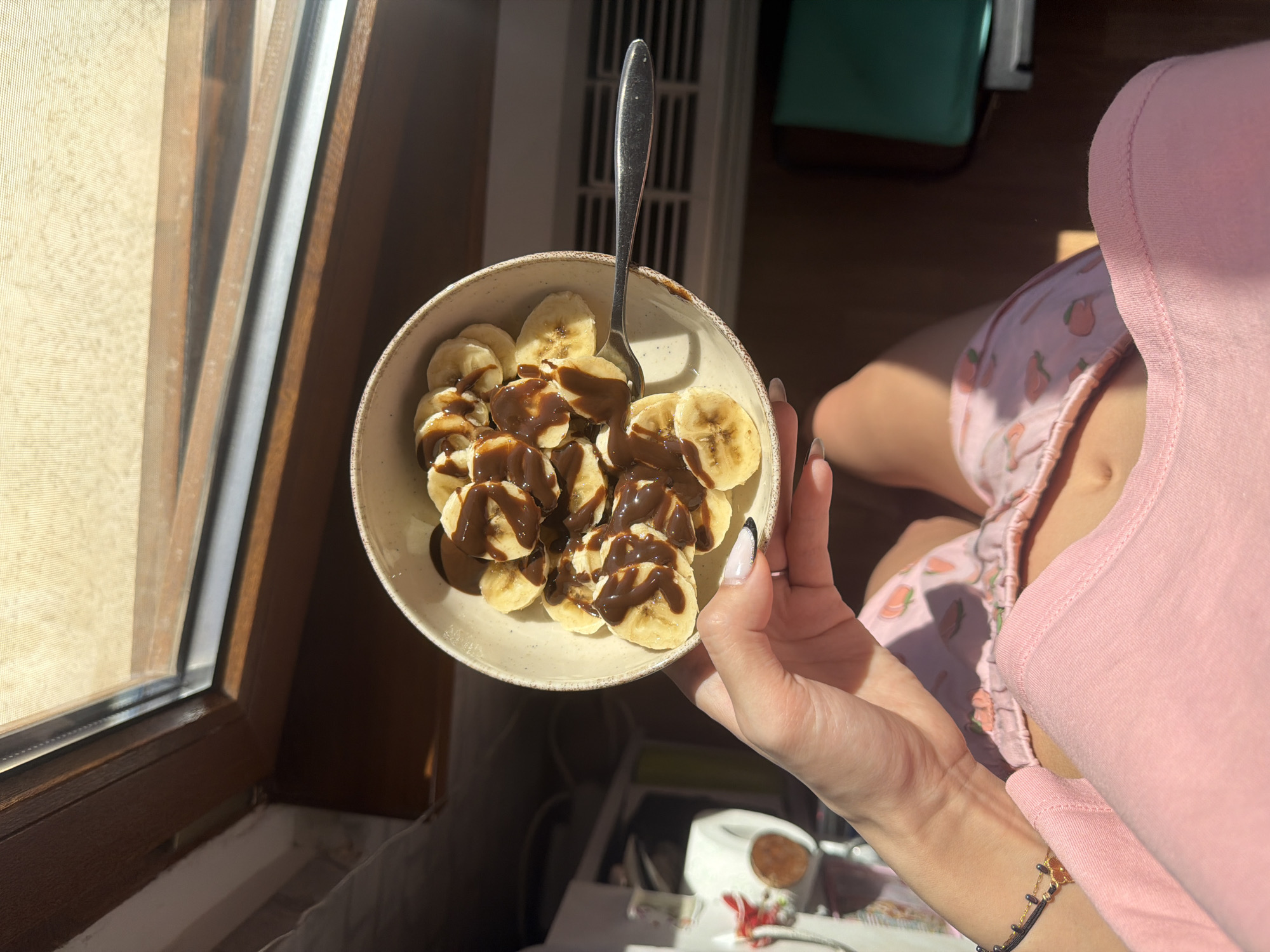 A person is holding a bowl of sliced bananas drizzled with chocolate sauce. A fork is in the bowl. The person is wearing a pink shirt and pink shorts with a strawberry pattern. The scene appears to be indoors, near a window. There is no identifiable information indicating the location in the city of Kyustendil, Bulgaria.