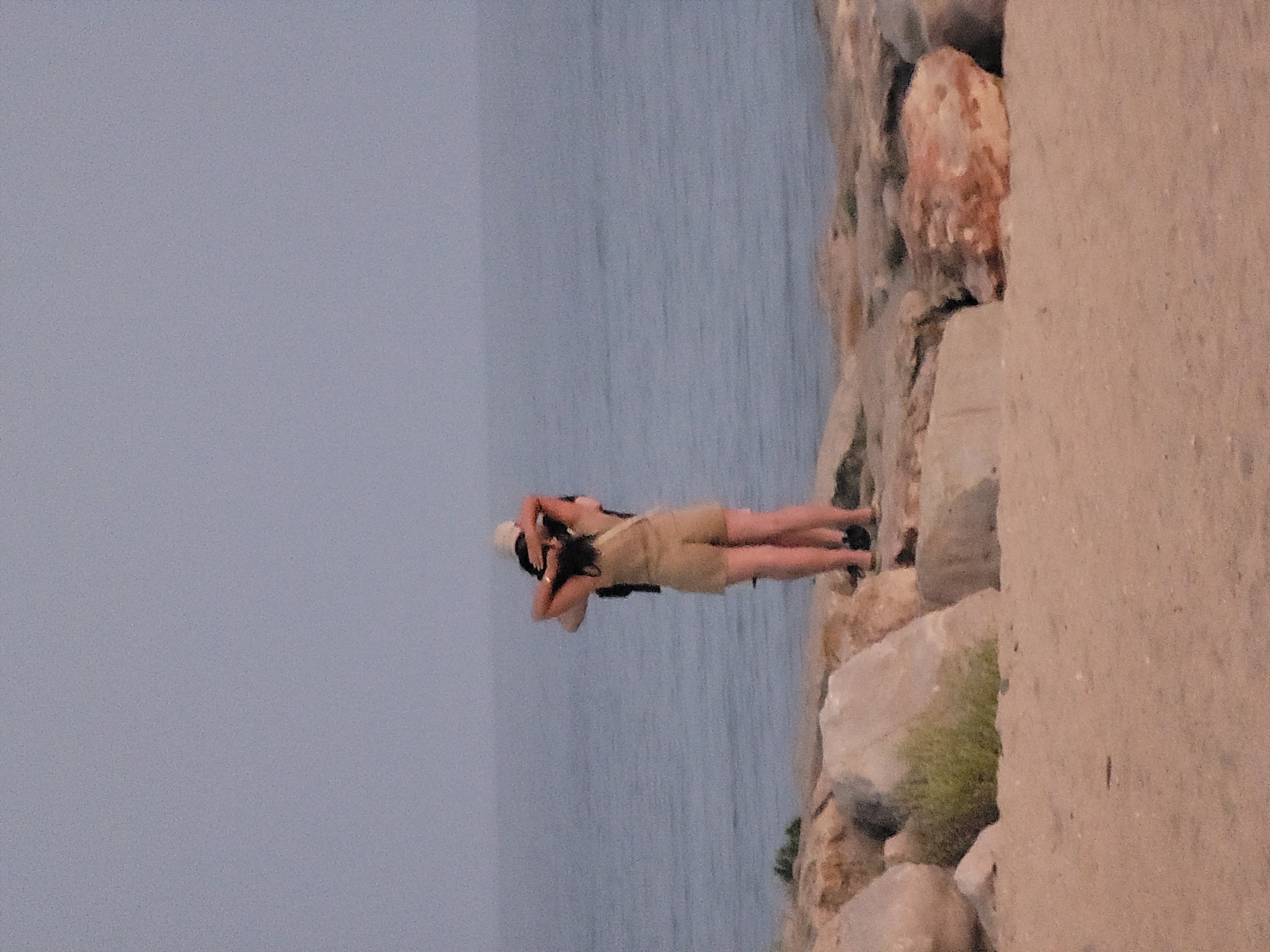 The image depicts two individuals embracing on a rocky shoreline in Quarteira, Portugal. The person in front, wearing a light-colored romper and a white cap, has their arms around the neck of the person behind them. The individual behind, wearing a dark top, has their arms wrapped around the other's shoulders. They are facing away from the camera, looking out at the ocean. The environment is characterized by a sandy beach in the foreground, a rocky embankment with sparse vegetation in the mid-ground, and a calm, overcast sea stretching to the horizon under a pale sky. The lighting suggests it is either early morning or late evening.