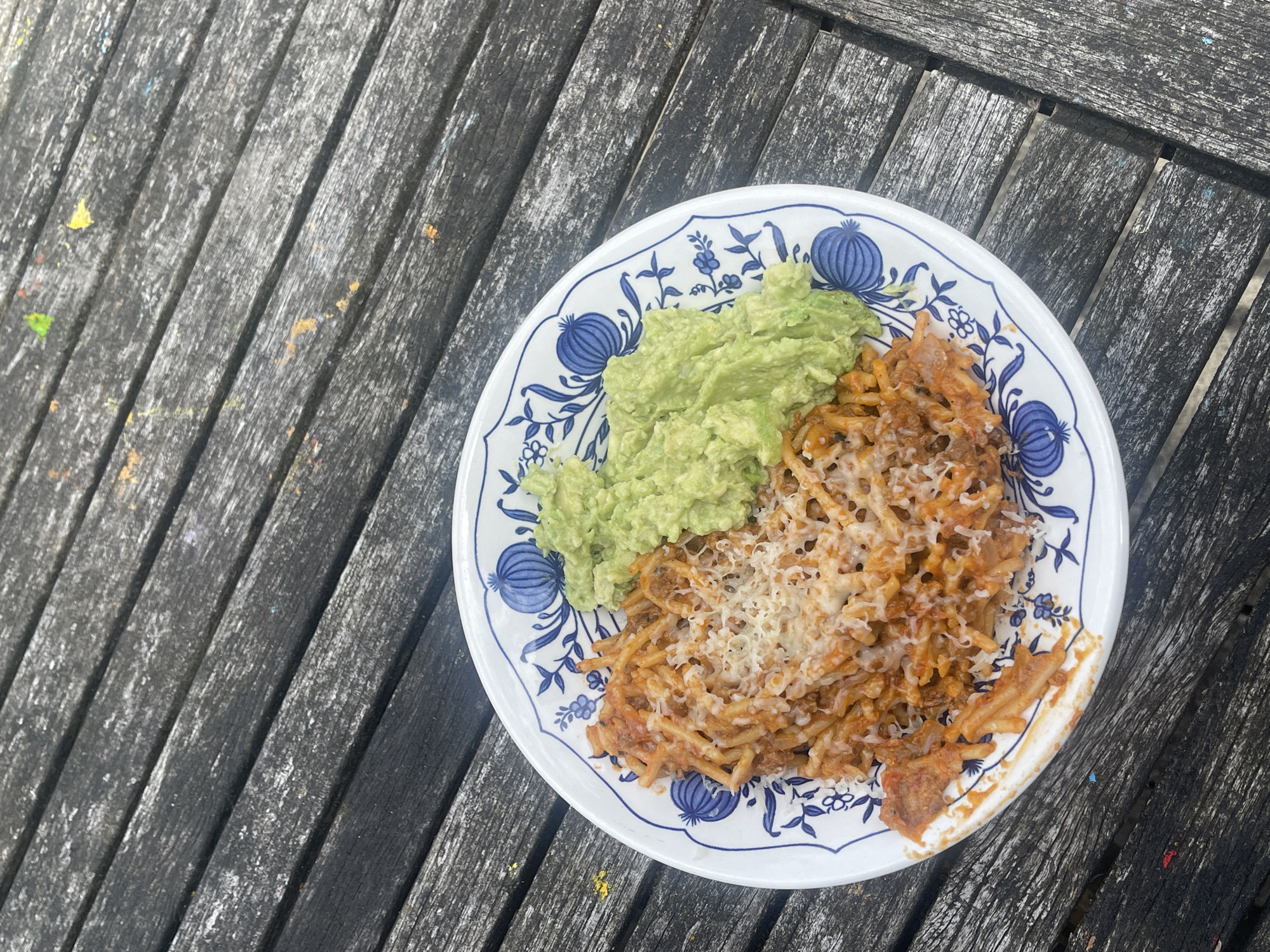 A plate of pasta and guacamole sits on a weathered wooden table. The plate has a blue and white floral pattern. The pasta is covered in a red sauce and topped with shredded cheese. The guacamole is to the right of the pasta. The wooden table is outdoors and shows signs of age and wear, with visible grain and some scattered paint splatters. The scene is captured from a top-down perspective. The location is not specified in Maidstone, United Kingdom.