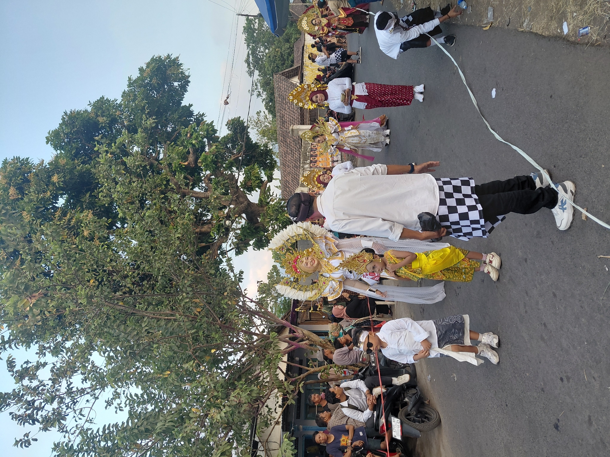 The image captures an outdoor cultural procession on a street in Bimo, Indonesia. A large, ornate costumed figure, likely a dancer or performer, stands prominently in the center, adorned with a golden headdress and white winged embellishments. To her left, a young child dressed in a yellow traditional outfit and headdress stands, observing the scene. Several other performers in similar elaborate golden headdresses and traditional attire, predominantly white and red, are visible marching or standing in procession behind the central figure. A man in a white shirt and black and white checkered sarong stands facing away from the camera, looking towards the procession. Spectators line the sides of the street, with some sitting on motorcycles and others standing. The environment features a paved road, buildings with visible roofs in the background, and mature trees providing shade. The sky is a clear blue, indicating daytime. A man in a white shirt and black pants is seen bending down at the right side of the road, appearing to pick up trash. A white ribbon or streamer runs diagonally across the road.