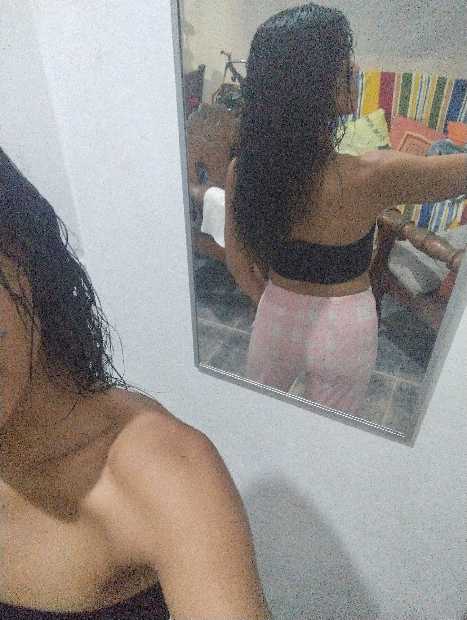 A person with long dark hair is facing away from the camera in front of a mirror. The person is wearing a black top and pink patterned pajama pants. The reflection in the mirror shows a room with a chair, striped pillows, and what appears to be a bicycle in the background. The room appears to be in a residential setting. The location, El Valle del Espíritu Santo, Venezuela, is not evident from the image.