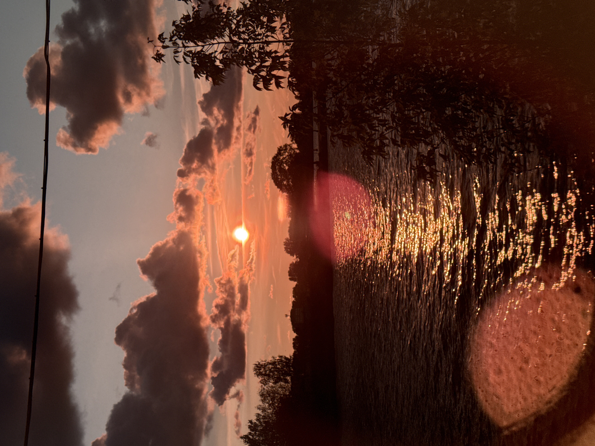 The image depicts a sunset over a body of water, likely a river or lake, with a silhouetted treeline along the far bank. The sky is filled with dramatic, reddish-orange clouds illuminated by the setting sun, which is a bright, small orb positioned slightly left of the center of the frame. The water reflects the vibrant colors of the sky, creating a shimmering, golden path. In the foreground, on the right side of the image, a silhouetted tree with dark foliage partially obscures the view. On the left side of the foreground, a large, reddish-pink, out-of-focus circular object is present, possibly a lens flare or a reflection. A dark, thin horizontal line, likely a power line, crosses the upper portion of the sky. The scene appears to be tranquil and natural. The provided context suggests this scene is in Riga, Latvia.