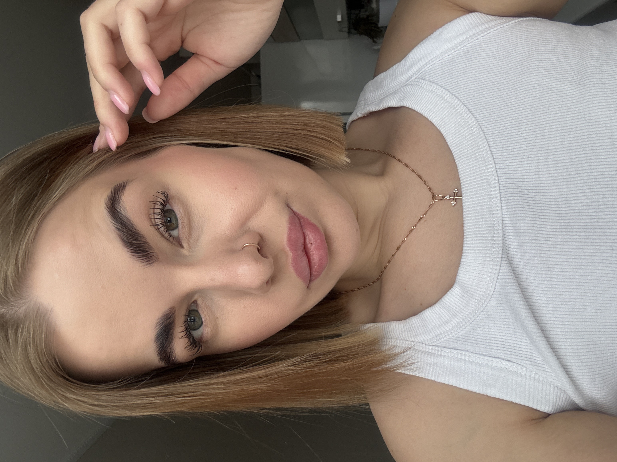 A selfie features a fair-skinned woman with light brown hair, wearing a white ribbed tank top and a delicate gold necklace with a small cross pendant. She has a nose ring in her left nostril and her left hand is raised above her head. The focus is on her face, which is angled slightly. It is not possible to confirm any location in the city of Chaikovsky, Russia, based on the image provided.