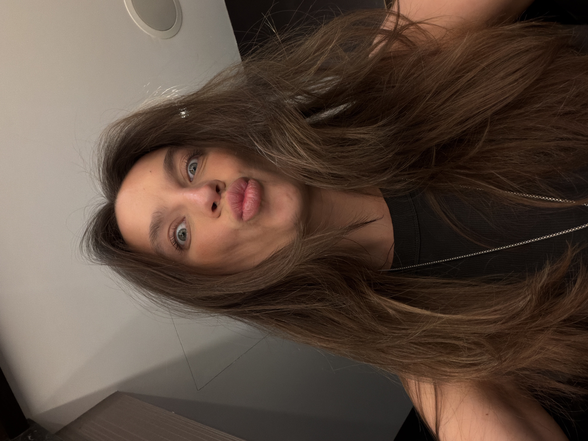 A young woman with brown hair and light eyes is posing for a selfie. Her hair is styled loosely around her face and shoulders. She is wearing a black top and a silver chain. The background is a muted gray and black. The photo was taken indoors. There is no indication the photo was taken in Budva, Montenegro.
