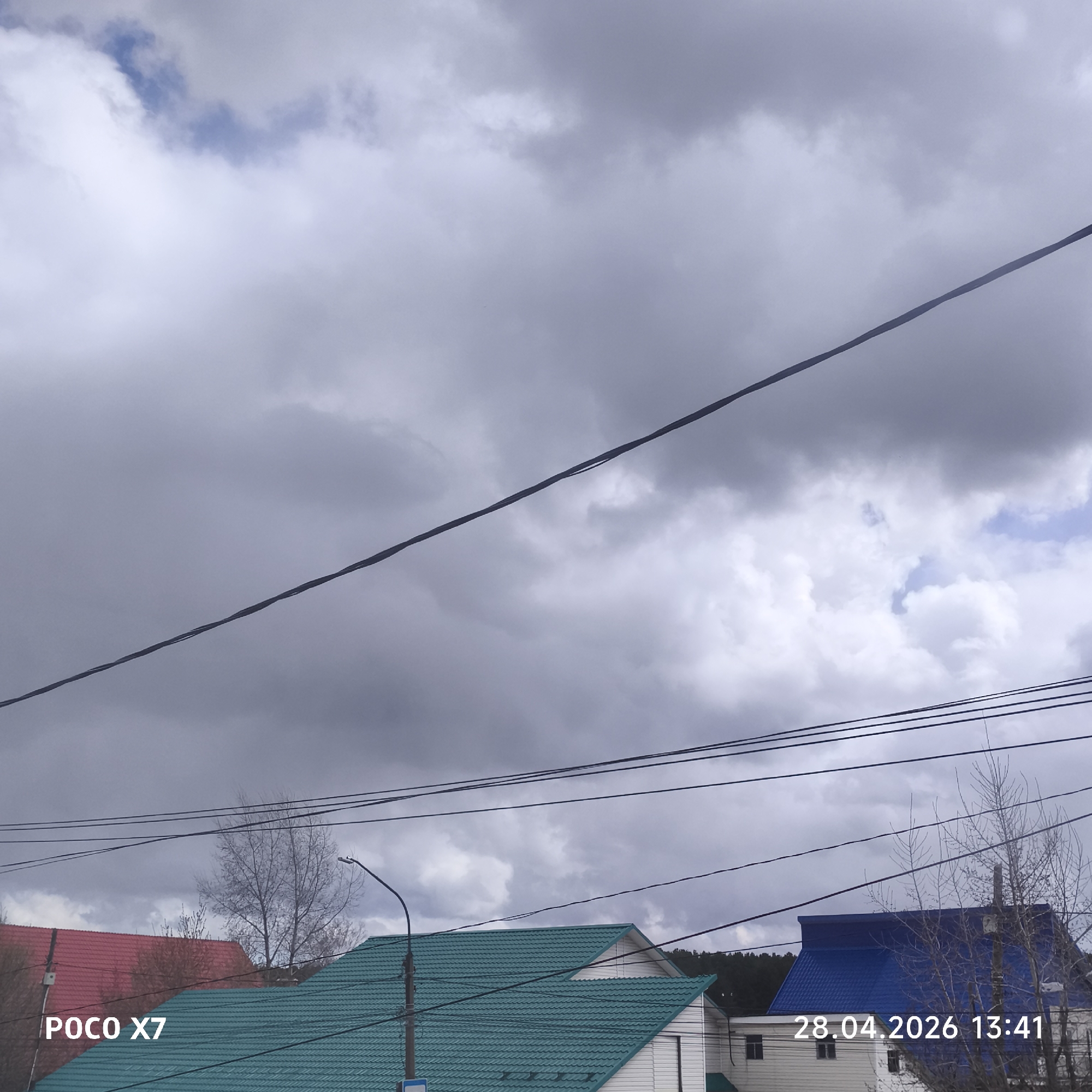 The image captures an outdoor scene, likely in a residential area of Chaikovsky, Russia, under a heavily clouded sky. The weather appears overcast with large, dense grey clouds dominating the upper and mid-sections, though patches of brighter white clouds and some blue sky are visible, particularly in the upper right.
Multiple dark power lines or cables stretch across the frame, some running horizontally in the mid-ground and one thick cable crossing diagonally from the upper left to the lower right. In the lower third of the image, three distinct houses are visible. On the far left is a house with a vibrant red, tiled roof. In the center, a house features a bright teal or green tiled roof and white siding. To the right, partly obscured by bare trees, is a house with a striking blue tiled roof and white siding.
Several deciduous trees, mostly bare of leaves, indicate either early spring or late autumn. A black streetlight with a curved arm stands to the left of the teal-roofed house. In the distant background, a dense line of darker trees suggests a forested area or hillside.
No specific activity or people are depicted. Visible text includes "POCO X7" in the bottom left corner, indicating the device used, and a timestamp "28.04.2026 13:41" in the bottom right, marking the date and early afternoon time.