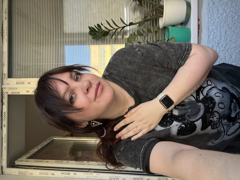 A female subject is depicted in a selfie-style, waist-up portrait taken indoors, within the city of Noviy, Russia. She has dark brown hair styled with bangs, light complexion, and is looking directly at the camera with a slight smile. Her hair is partially secured, revealing a lavender hair tie near her left ear, adjacent to a gold-toned triangular earring with a clear central stone. She wears a washed-out dark grey t-shirt featuring a sparkly black Mickey Mouse design, a thin gold-colored chain necklace, a gold-toned ring on the ring finger of her right hand, and a light-colored Apple Watch with a gold-toned frame on her left wrist. Her right hand is positioned on her left shoulder.
The background consists of a white PVC window frame with the repeated vertical text "GOODWIN IN". The left window pane is open inward, while the right pane is closed, displaying horizontal white blinds partially obstructing the external view. A white window sill runs beneath the closed pane, supporting two potted plants: a taller, dark green plant in a white pot and a smaller plant with reddish leaves in a teal-green pot. Beyond the window, a clear blue sky is visible above distant light-colored urban buildings.
