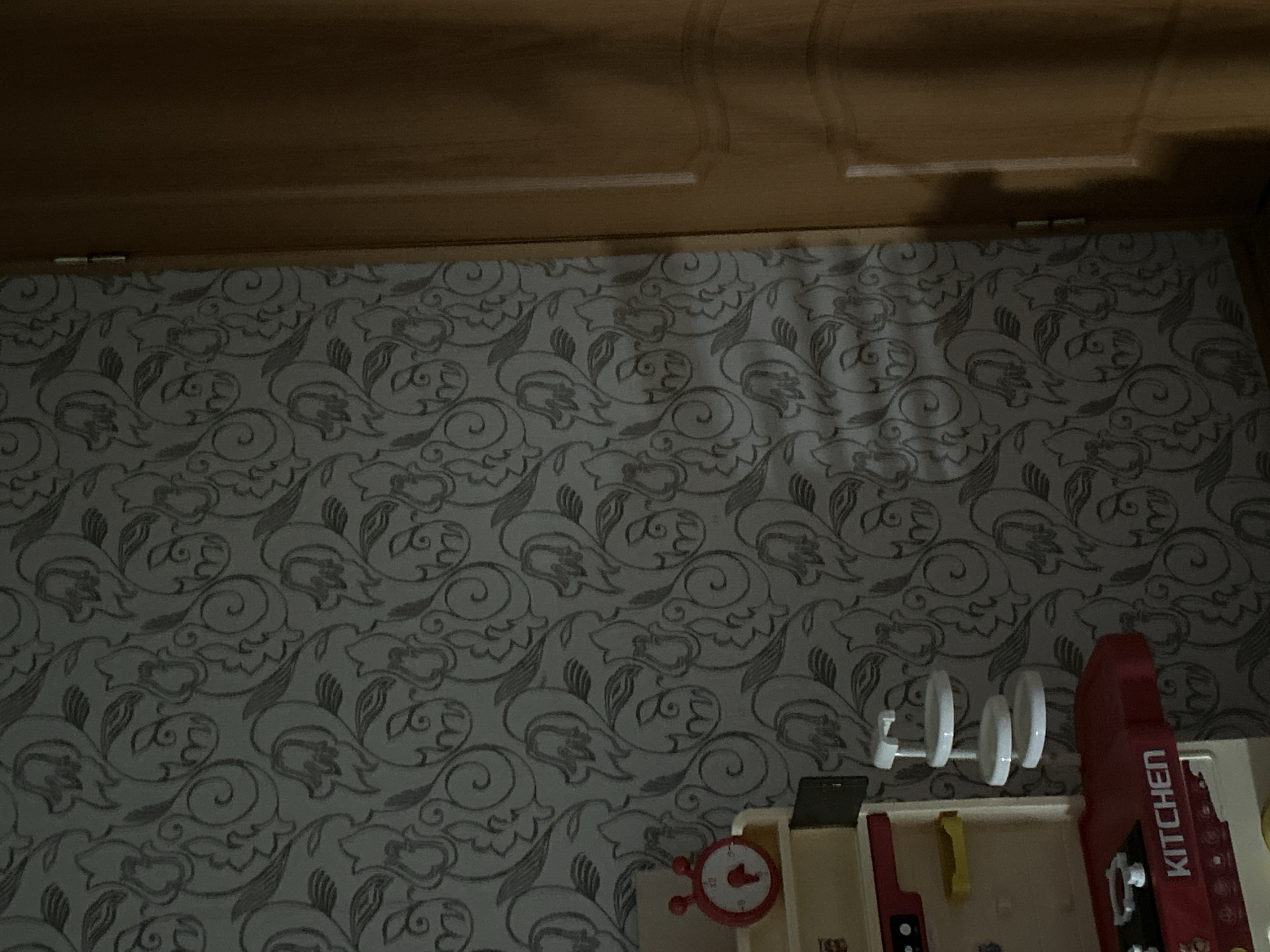This media file is a close-up, low-angle shot of a portion of a room, likely a child's play area. The foreground features a beige toy kitchen set. Visible on the toy kitchen is a red clock with white hands and red knobs. To the right of the clock is a red toy appliance with the word "KITCHEN" printed vertically in white letters. Next to the toy appliance, there is a white toy with three circular elements mounted on a white horizontal bar. Behind the toy kitchen, the wall is covered with wallpaper featuring a repeating pattern of stylized flowers and leaves in a light gray color. The top portion of the image shows the underside of a wooden surface, possibly a shelf or cabinet, with visible wood grain and a shadow cast across it. The lighting suggests it is daytime, with a shadow indicating a light source from above and to the left, creating a dimly lit environment. The focus is on the toys and the patterned wallpaper, with the wooden structure serving as a backdrop.