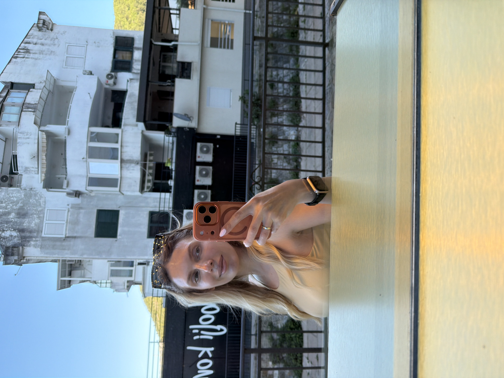 A young woman with long blonde hair, wearing a light-colored top, is captured in a selfie reflected in a yellow-toned, possibly wooden or laminated, surface in the foreground. She holds an orange-cased smartphone with her right hand, her left hand adorned with a ring and a smartwatch. Sunglasses are perched on her head, and she looks directly at the camera with a gentle smile.
The scene is an outdoor urban setting, likely a cafe or restaurant patio, in Budva, Montenegro. Behind the woman, visible through the reflection, are several multi-story white buildings with multiple windows, some fitted with dark shutters. Balconies and external air conditioning units are also visible on the building facades. A metal railing or fence separates the foreground from these buildings. In the upper background, a clear blue sky is visible, and further in the distance, a verdant mountain range rises.
The time of day appears to be bright daytime, with clear weather. A notable detail is a dark sign below the buildings displaying white, reversed text that appears as "KONJIJool", indicating a business name or branding. The image is taken through a reflective medium, causing the background elements, including the sign, to appear mirrored.