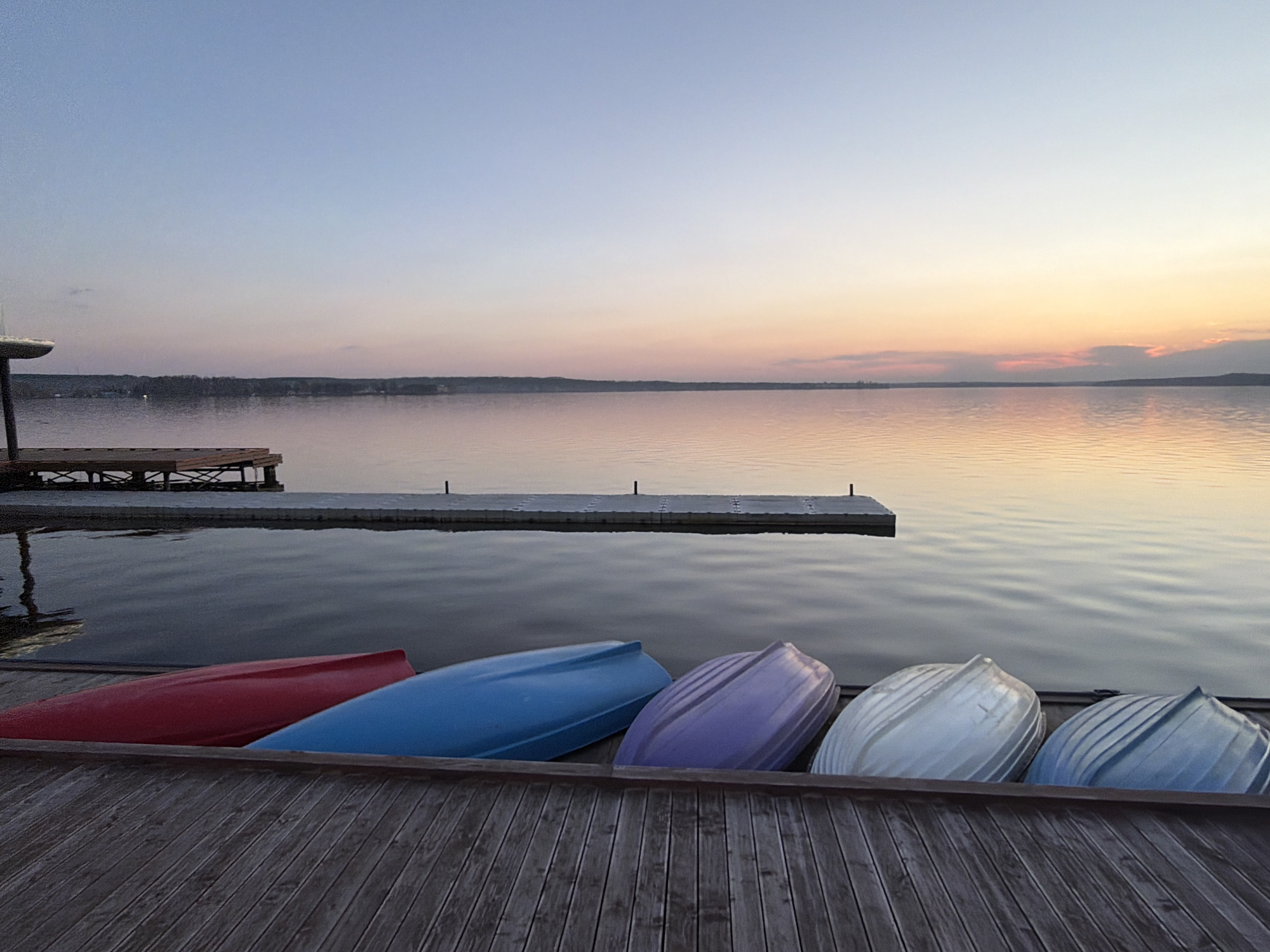 The image depicts a tranquil scene at dusk or dawn on a calm body of water, possibly a lake, with several colorful boats resting on a wooden dock. From left to right, the boats are colored red, blue, purple, white, and blue. A longer dock extends into the water to the left of the boats. The sky is a gradient of pale blues, pinks, and oranges, suggesting either sunrise or sunset. The water reflects the colors of the sky, creating a smooth, mirror-like surface. The horizon line shows trees in the background, indicating land on the opposite side of the water. There are no people visible in the image.
The image is of Lake Votkinsk, at a boat launch in Votkinsk, Russia.