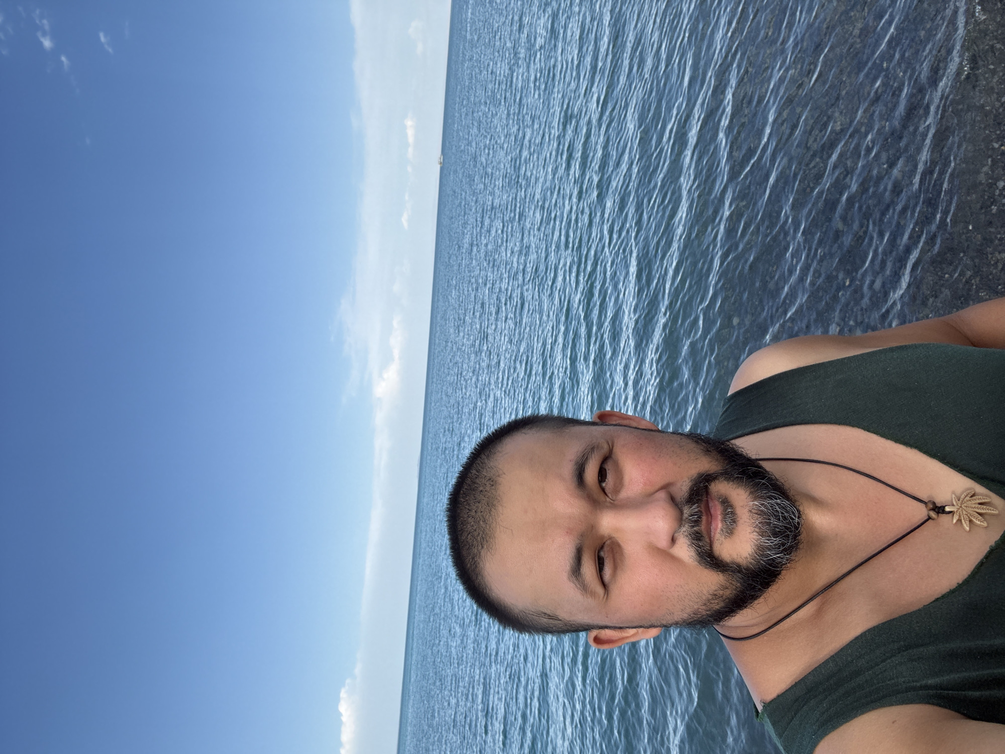 A man with a beard and short hair is taking a selfie by the water. He is wearing a dark green sleeveless shirt and a necklace with a pendant. The background shows a blue sky with some clouds and a vast body of water. The image appears to be taken outdoors, possibly at a lake or sea, in the vicinity of Ақши, Kazakhstan. The water is calm with small ripples, and the seabed is visible in some areas. There is a small boat in the distance on the horizon.