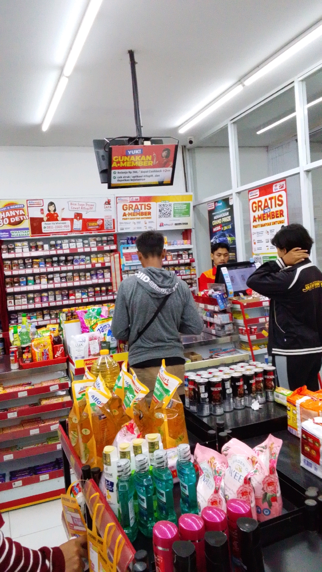 The image depicts the interior of a convenience store, likely an Alfamart, in Desa Karangbenda, Indonesia. The store is well-lit with fluorescent ceiling lights. Shelves are stocked with various products, including snacks, drinks, personal care items like mouthwash and shampoo, and cooking oil. In the foreground, there are displays of bottled oil, packaged snacks, and personal hygiene products.
Three individuals are visible. One person, wearing a gray hoodie and black pants, is standing facing away from the camera, appearing to browse the shelves. Another person, in a black jacket with white lettering and black pants, is standing to the right and slightly behind the first person, with their hand touching their neck. A third person, wearing a red and yellow uniform, is behind a counter to the right, looking at a computer screen. There is also a digital screen hanging from the ceiling displaying promotional material for "YUKI GUNAKAN A-MEMBER". The store has large windows to the right, suggesting it's a street-facing location.