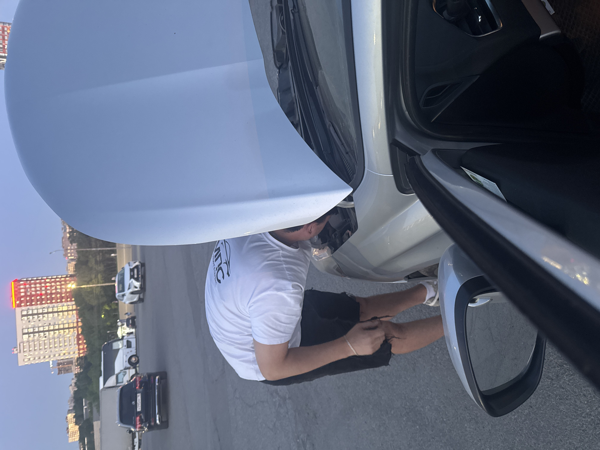 The image shows a man in a white t-shirt and dark shorts inspecting the engine of a silver car with the hood open. The driver's side door is also open. The scene appears to be a parking lot or roadside in the city of Izhevsk, Russia. In the background, there are several cars parked and a tall, multi-story building with red accents on the upper floors. The sky is dusky, suggesting either early morning or late evening. The pavement is asphalt.